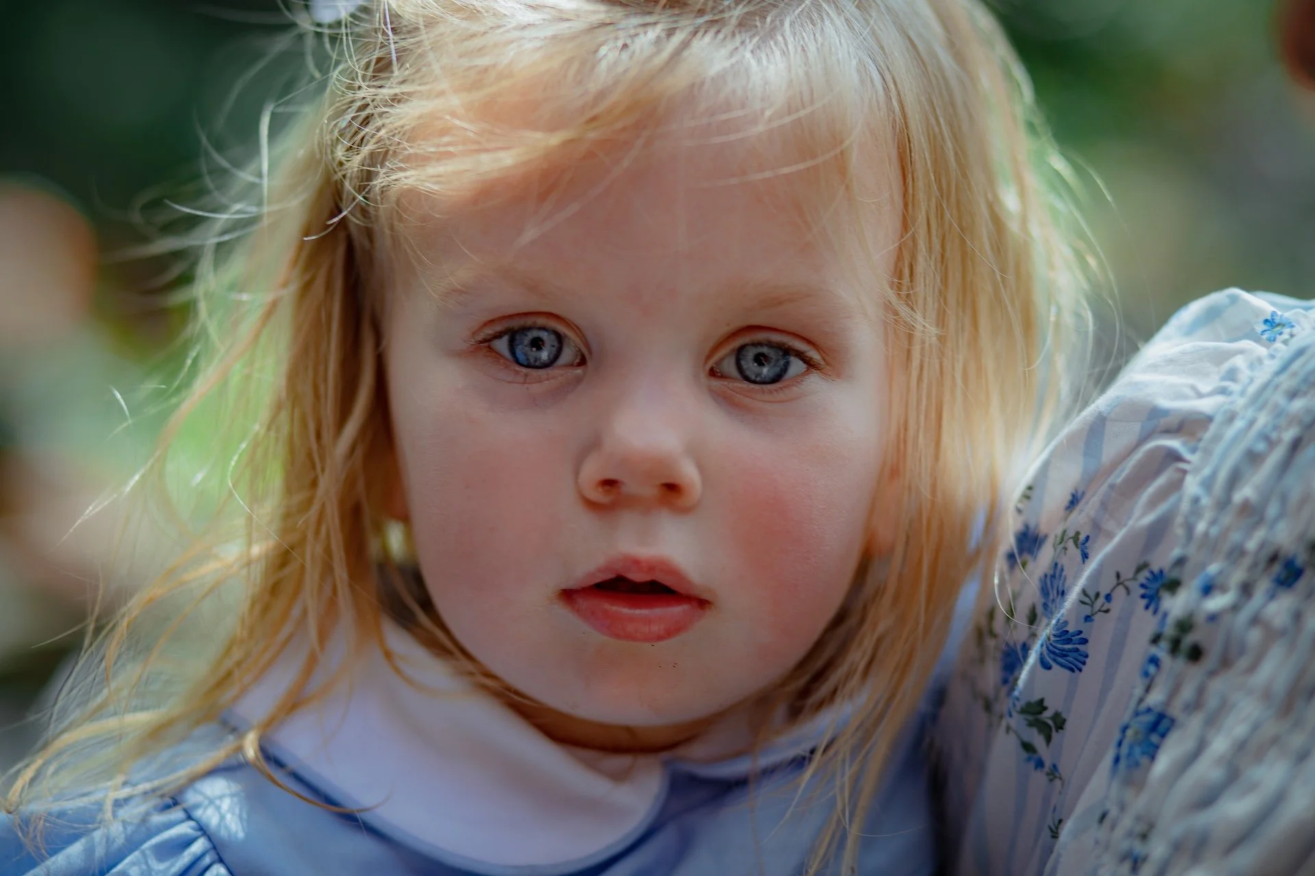 Close-up of a young girl with blue eyes and light blonde hair outdoors, wearing a white and blue floral dress. L'objectif Photography: New Orleans Wedding, Event, and Portrait Photographer
