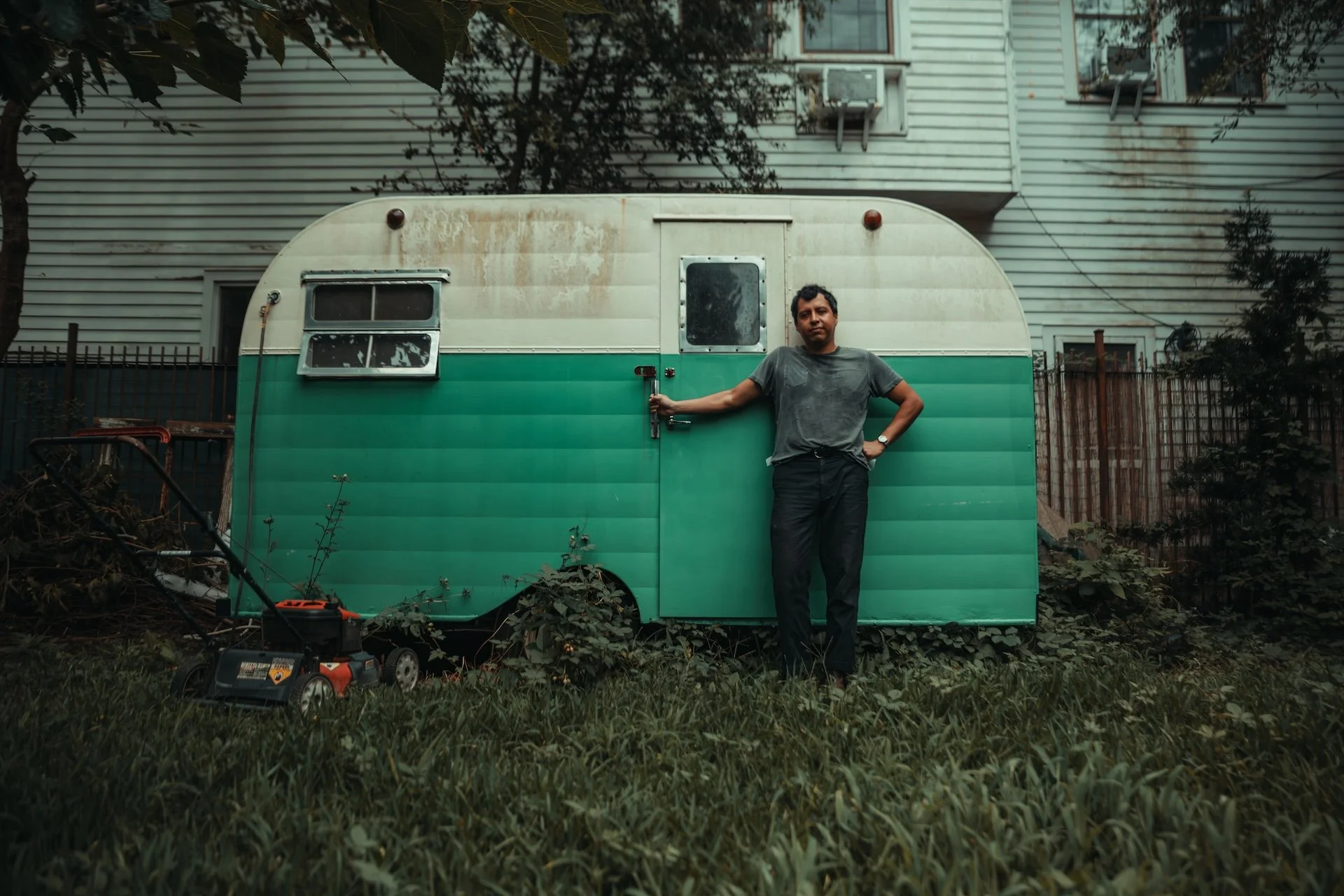 A man standing outdoors next to a vintage green and white trailer with his hand on the door handle, surrounded by grass, trees, and a house with worn siding in the background. L'objectif Photography: New Orleans Wedding, Event, and Portrait Photograp