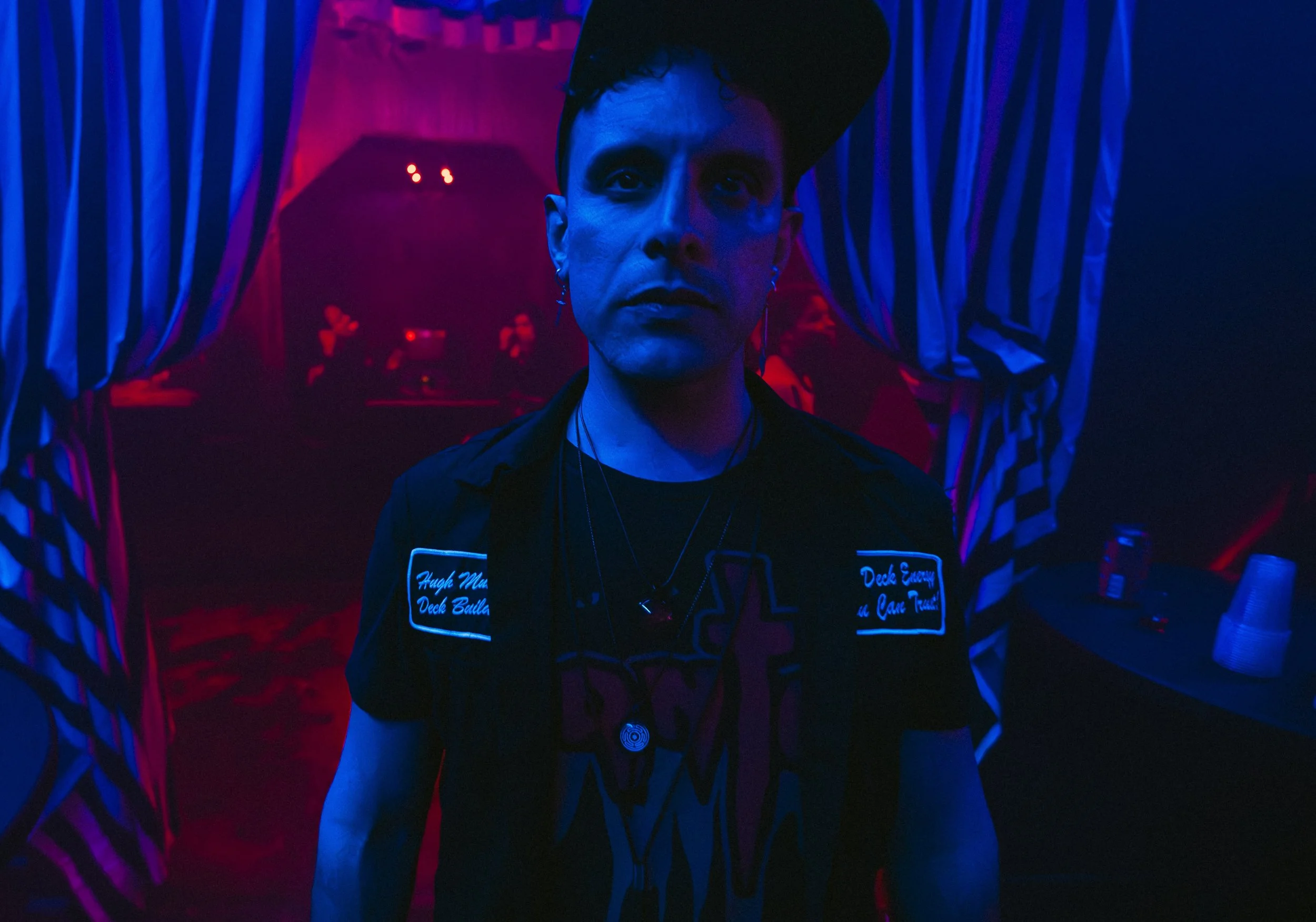 A person with short dark hair and earrings, illuminated by blue and red lighting, standing in front of striped curtains with a red backdrop and some people in the background. L'objectif Photography: New Orleans Wedding, Event, and Portrait Photograph