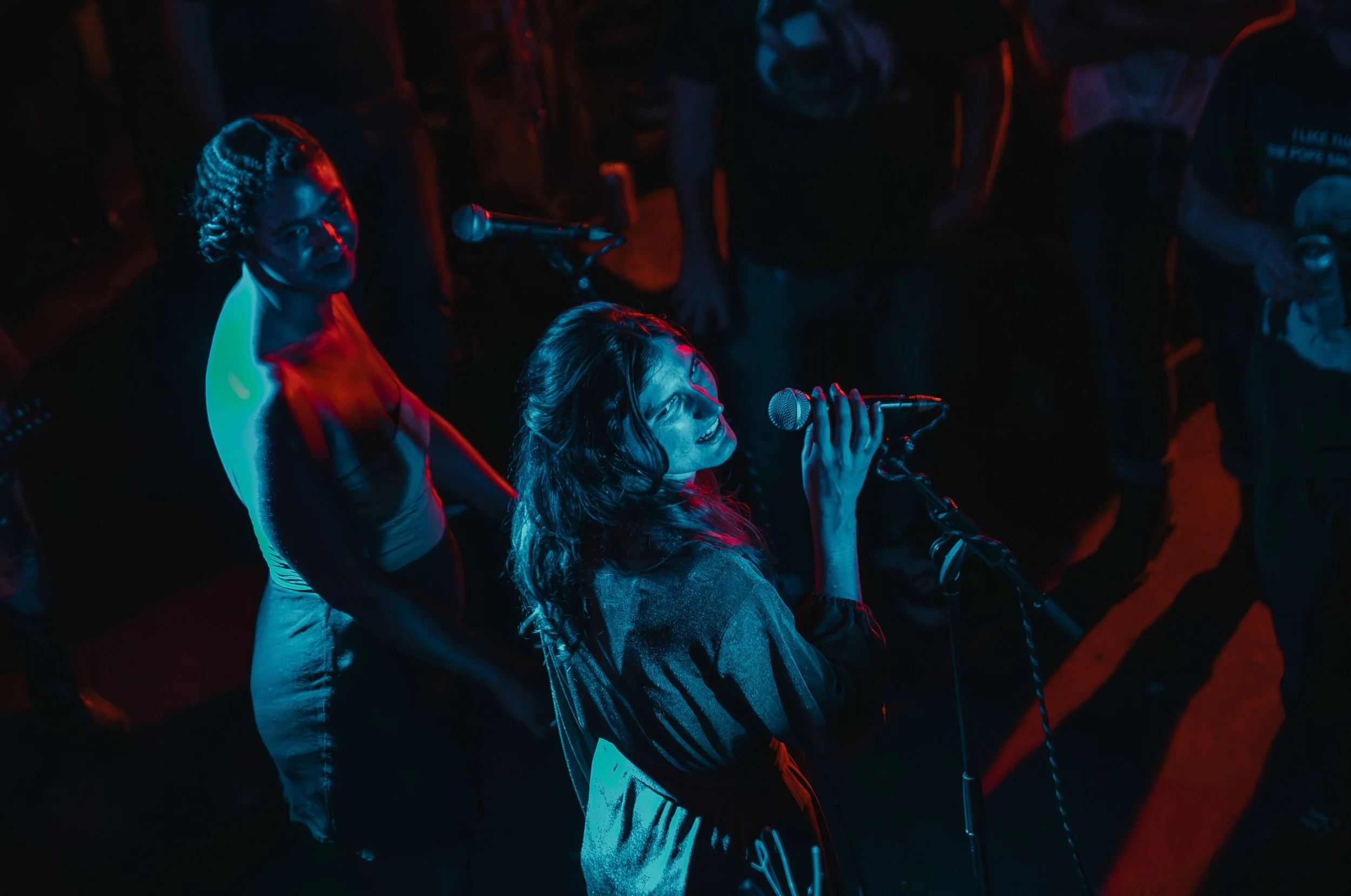 Woman singing into a microphone at a music event, with people surrounding her in a dark environment illuminated by blue and red lights. L'objectif Photography: New Orleans Wedding, Event, and Portrait Photographer