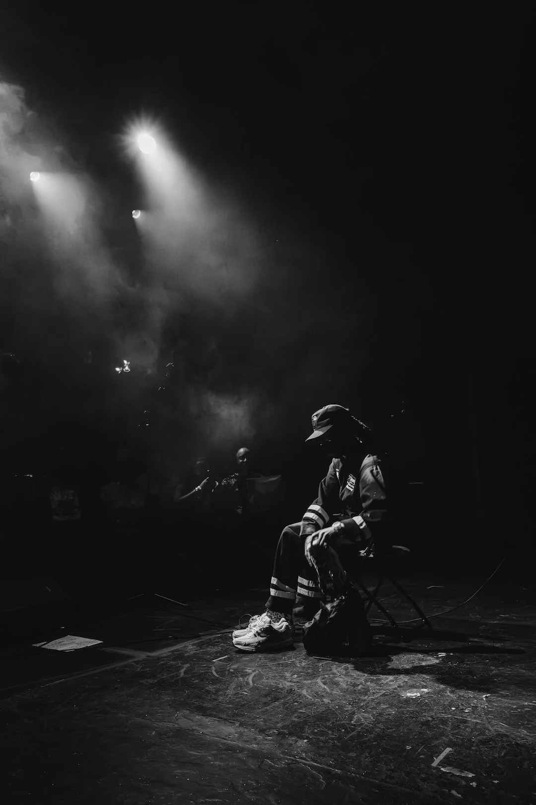 A person wearing a cap and casual clothing, sitting on a small chair on a dimly lit stage with smoke and spotlights overhead, with other people faintly visible in the background.