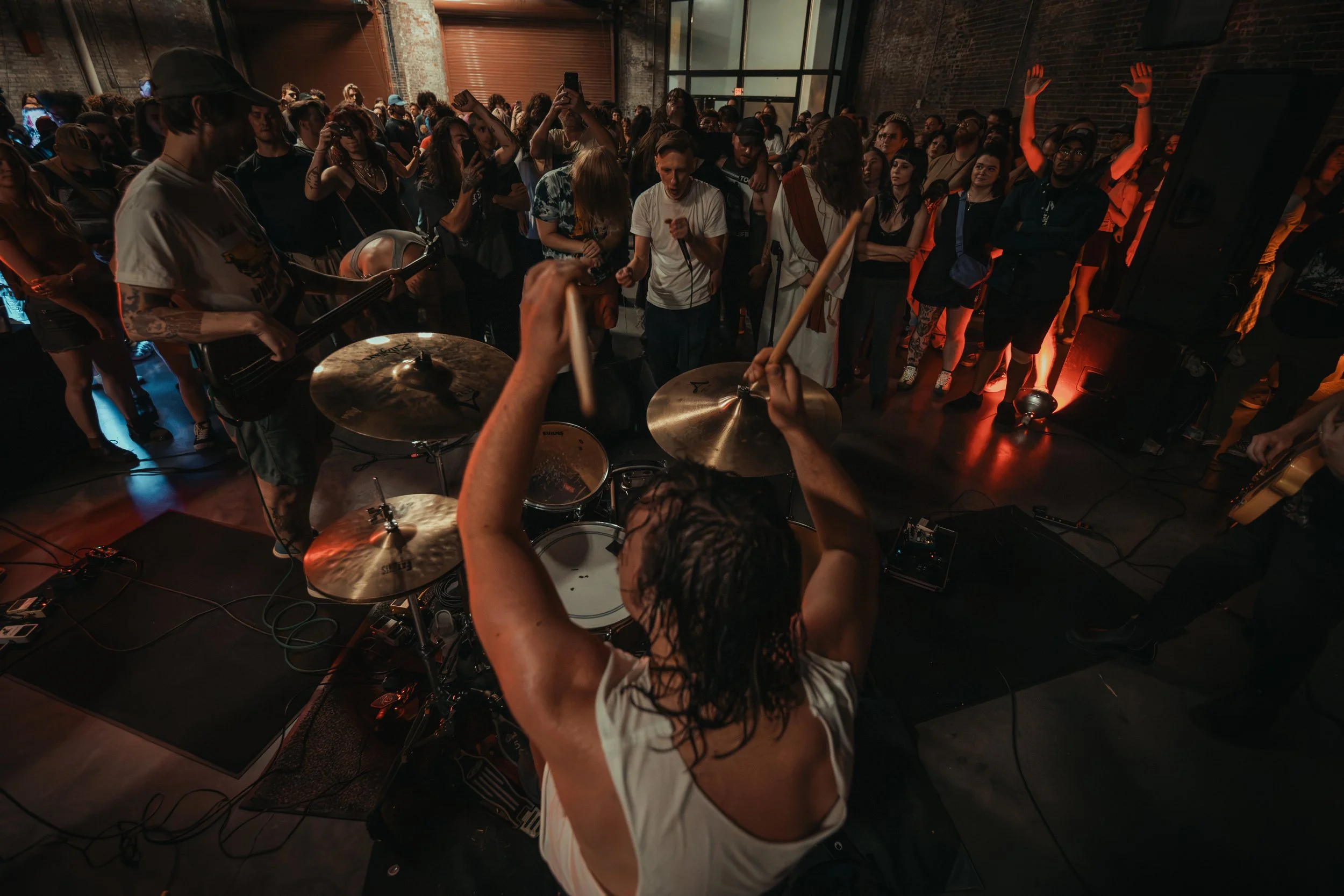 A group of people at a live music event, with the focus on a drummer and a guitarist performing on stage, surrounded by an audience in an indoor venue with brick walls and industrial windows.