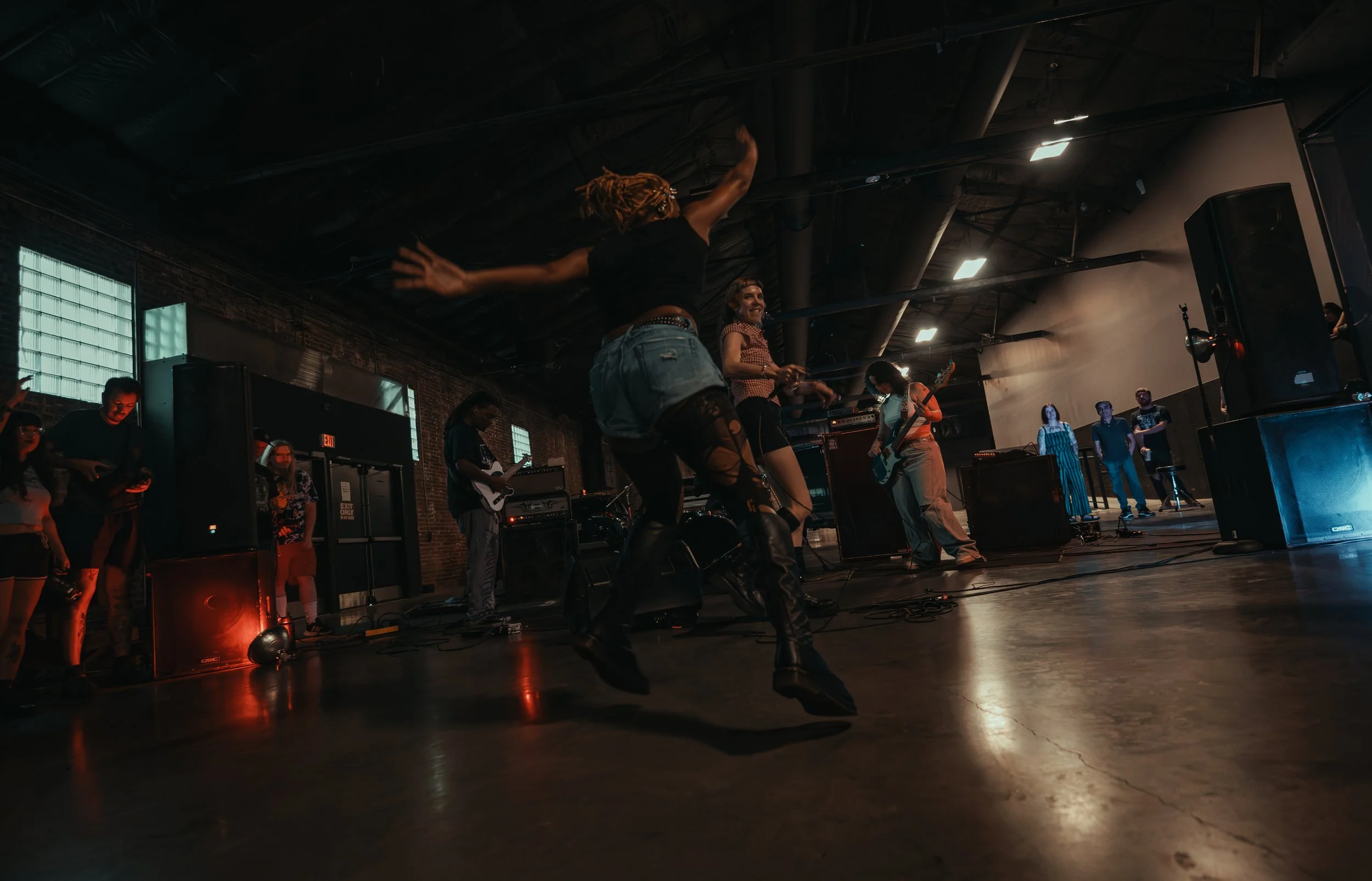 A live music performance with musicians playing guitars and singing on a stage inside a dark industrial-style venue, with some audience members visible in the background.