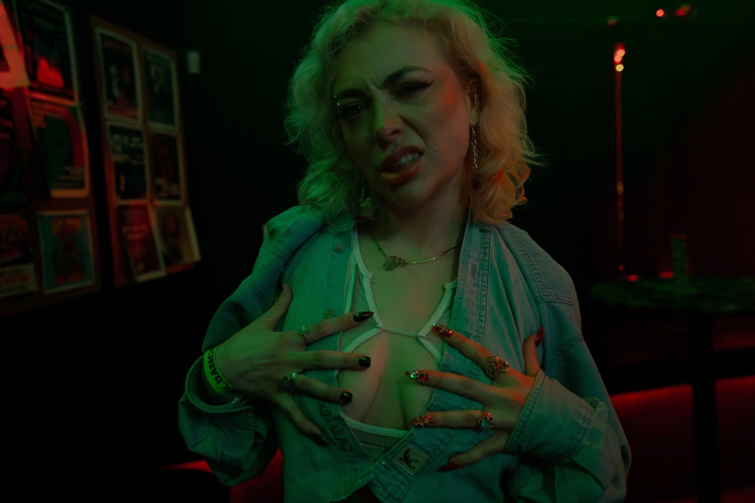 A woman with platinum blonde curly hair posing with a playful and defiant expression in a dimly lit room with neon green and red lighting. She is wearing a denim jacket, multiple rings, bracelets, and earrings, with her hands on her chest. L'objectif
