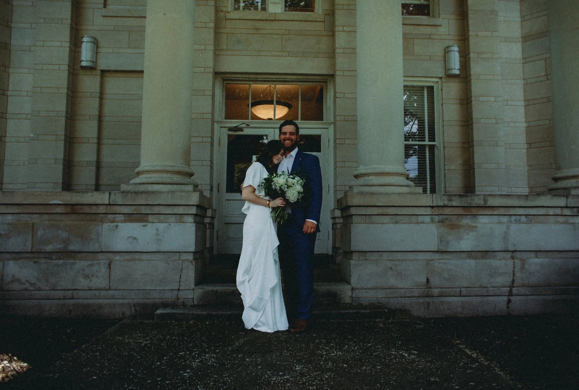 A bride and groom standing together on the steps of a large stone building, smiling. The bride is holding a bouquet of white flowers, and the groom is wearing a blue suit. L'objectif Photography: New Orleans Wedding, Event, and Portrait Photographer