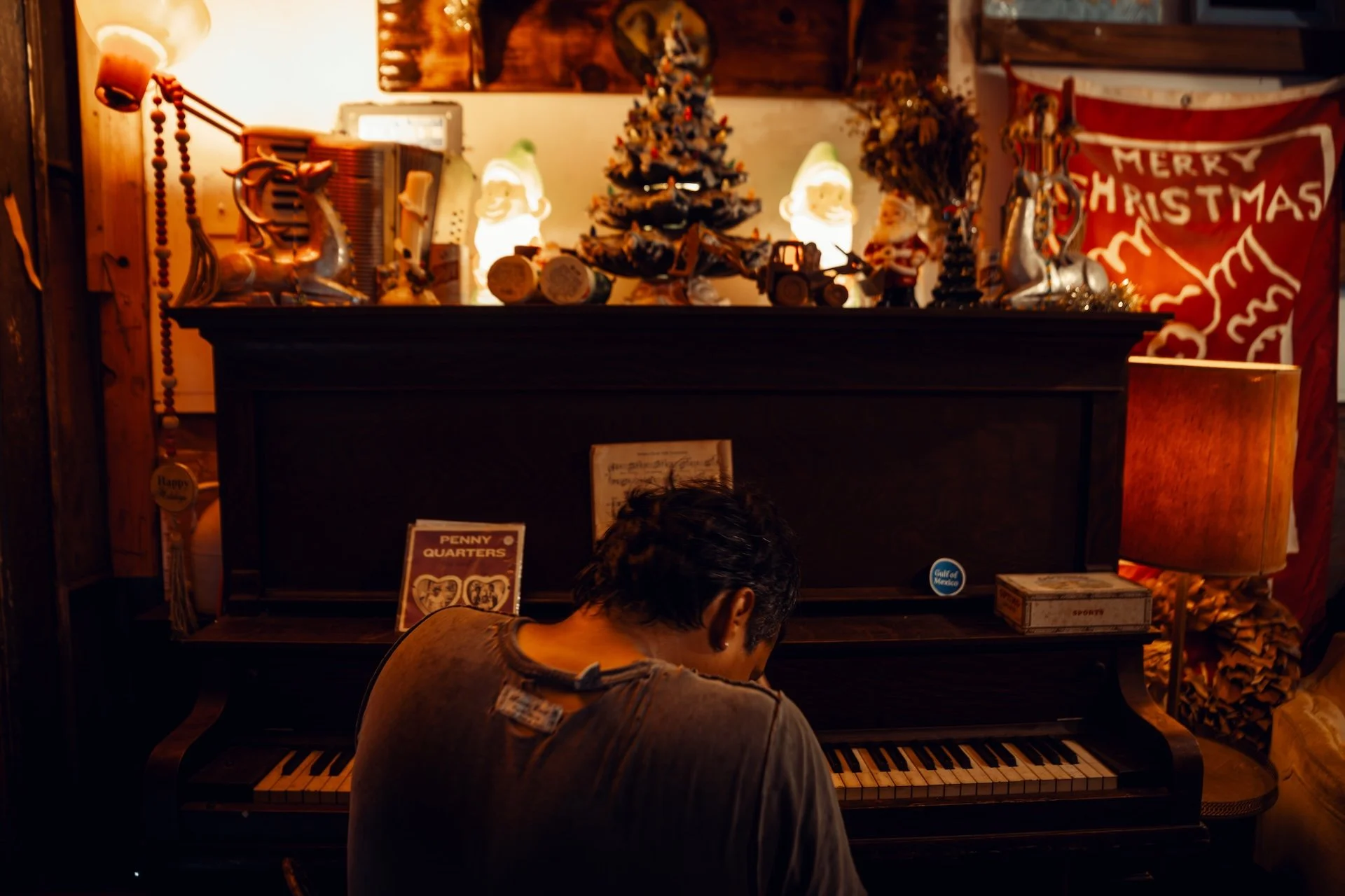 A person sitting at a black upright piano in a cozy room decorated with Christmas ornaments, including a small Christmas tree, ceramic elf figures, and a red banner with 'Merry Christmas' written on it, with warm ambient lighting. L'objectif Photogra