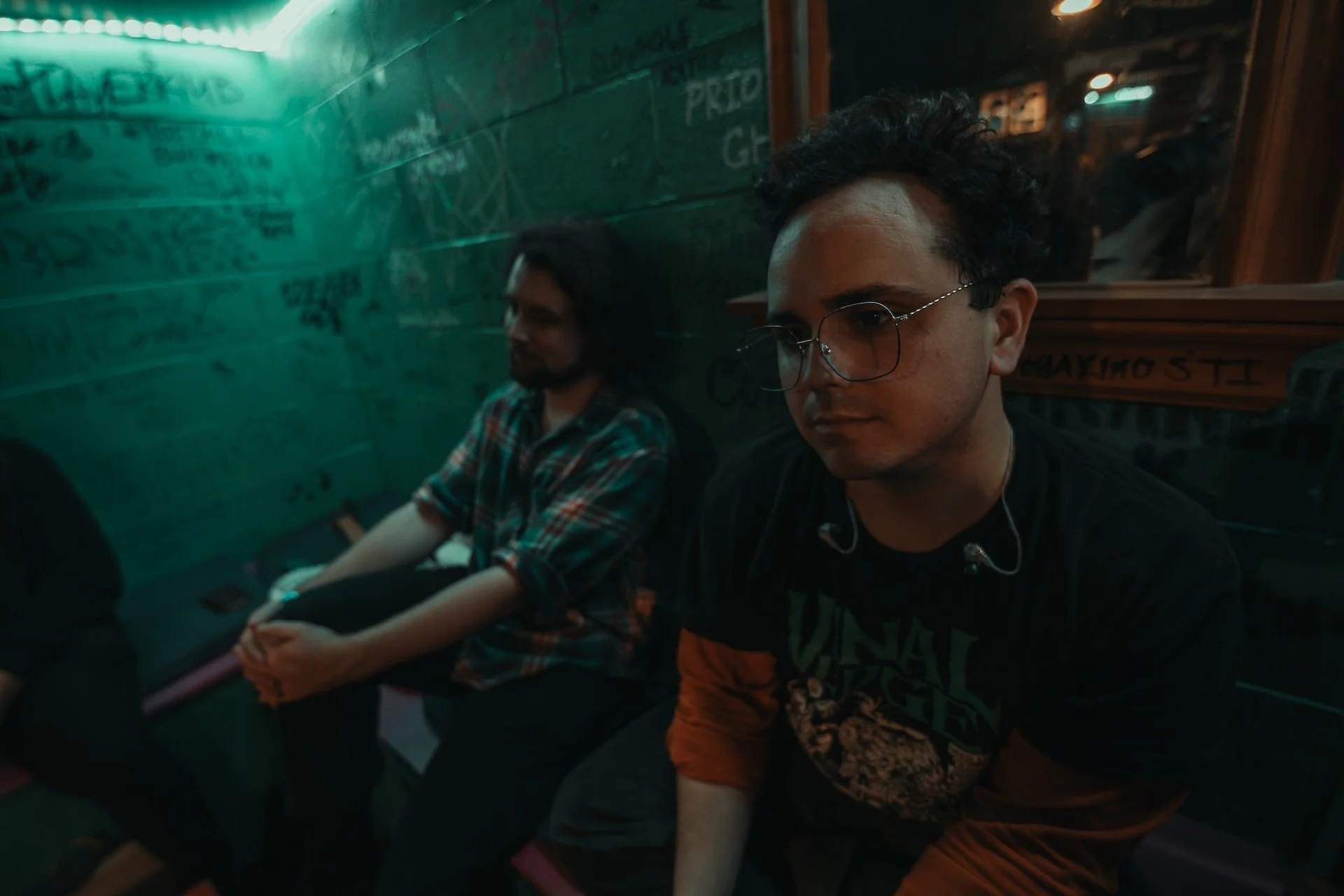 Two young men sitting inside a dimly lit restaurant or bar. The one on the right has curly dark hair, glasses, and is wearing a graphic T-shirt. The other has straight dark hair, a beard, and is wearing a plaid shirt. They are seated against a wall w