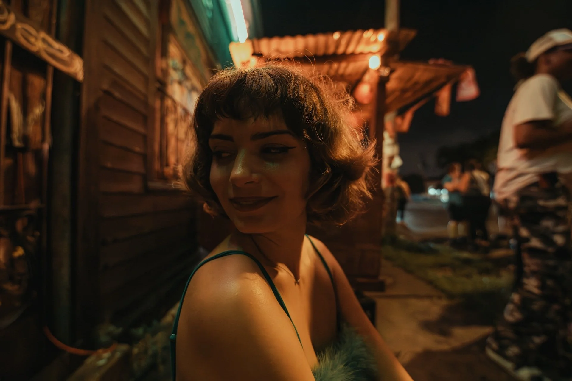 A woman with short curly hair and dark eyebrows is smiling and looking to the side at night, sitting outdoors near a wooden building with warm lighting, while people are in the background. L'objectif Photography: New Orleans Wedding, Event, and Portr
