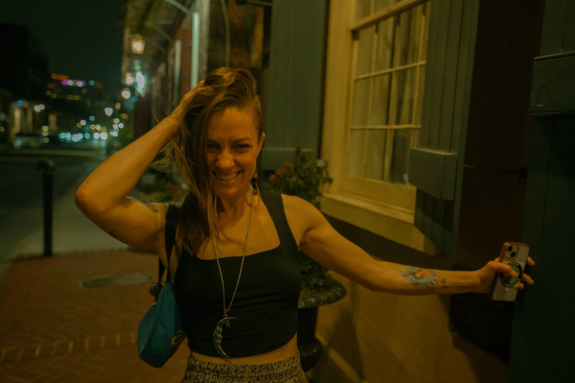 A smiling woman with long hair wearing a black tank top and a moon necklace taking a selfie at night on a sidewalk outside a building with wooden siding and a window, in an urban area with streetlights and blurred vehicle lights in the background. L'