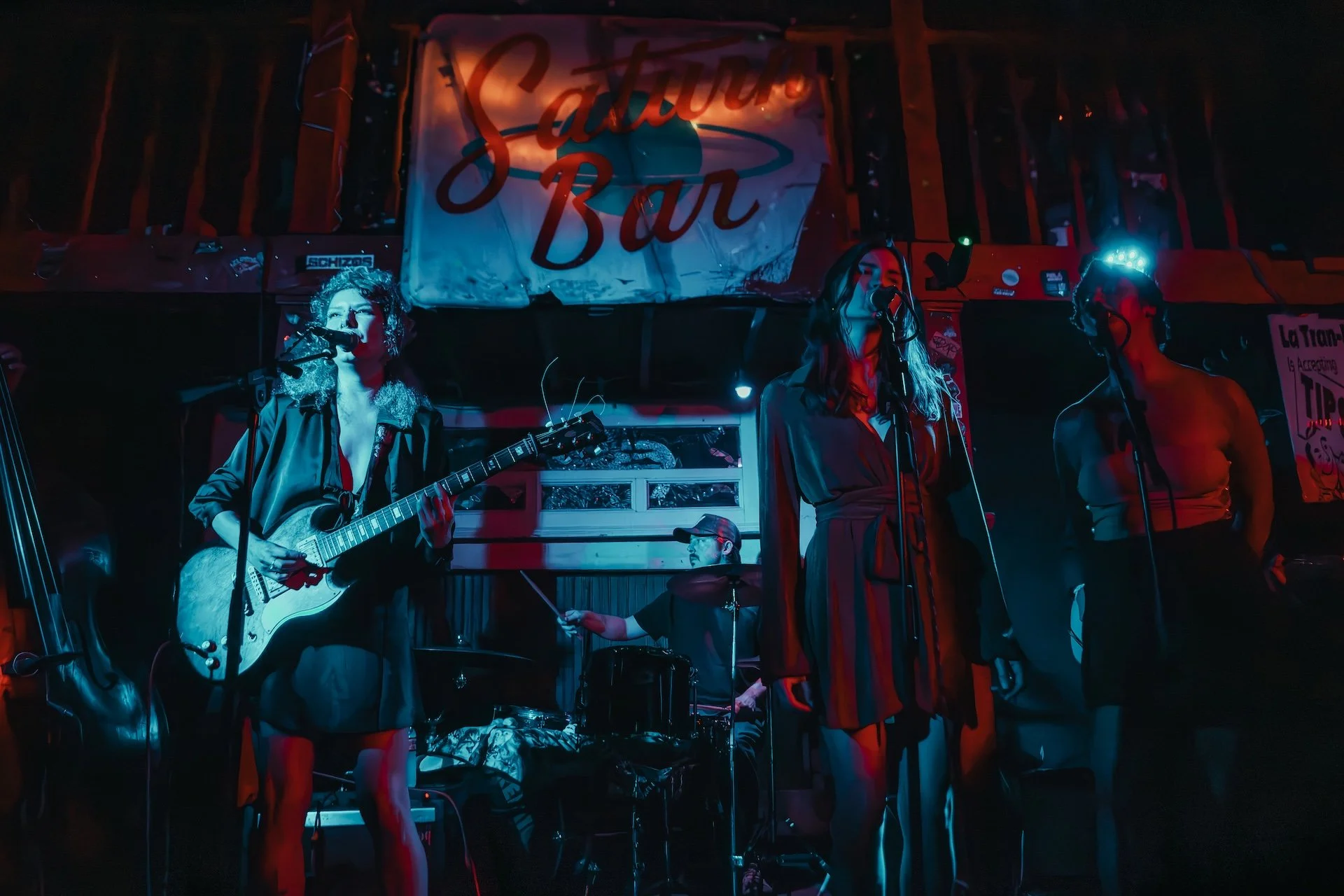A band performs on stage at a venue with a red and white sign that reads 'Saloon Bar.' The scene is illuminated with blue and red stage lights, showing a female guitarist, two female singers, a drummer, and a male musician, surrounded by musical equi