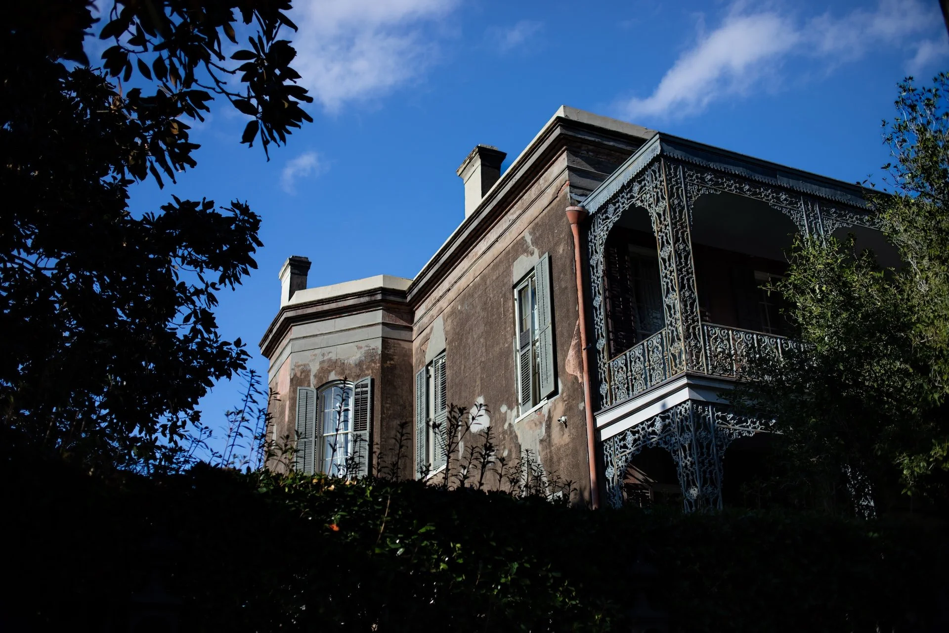 A multi-story brick building with a decorative metal balcony on the second floor, surrounded by trees, with blue sky and clouds overhead. L'objectif Photography: New Orleans Wedding, Event, and Portrait Photographer