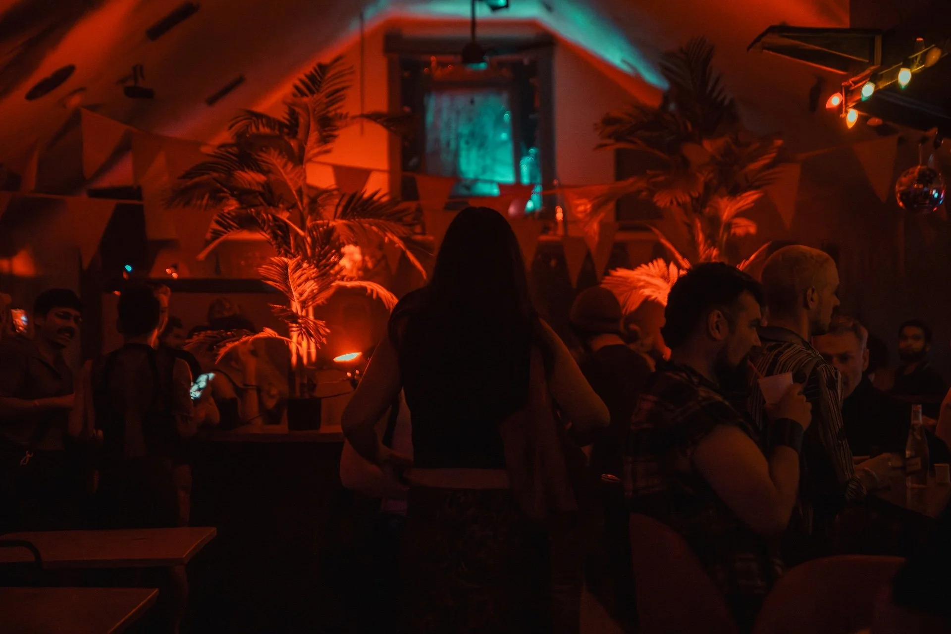 Dark indoor party scene with people standing and sitting, colorful ambient lighting, palm plants, bunting decorations, and a disco ball. L'objectif Photography: New Orleans Wedding, Event, and Portrait Photographer