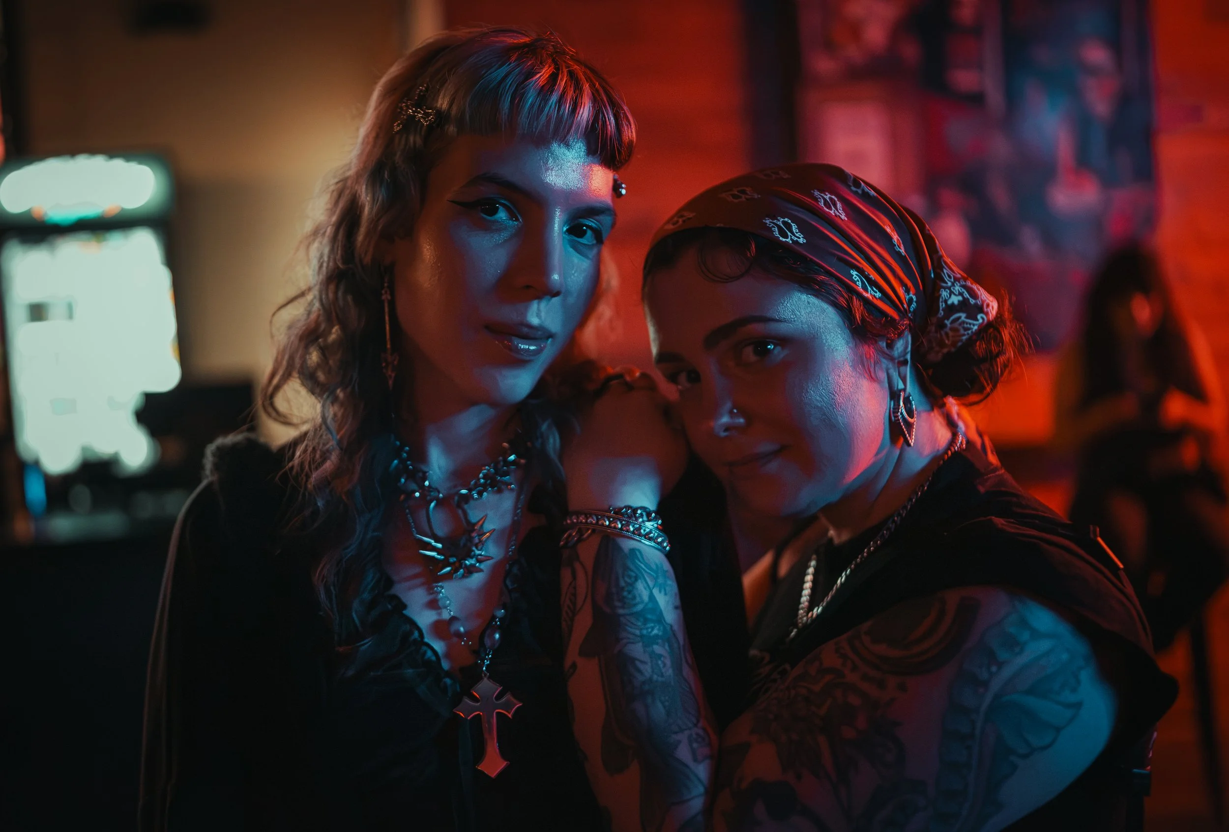 Two women with bold makeup and jewelry, one with short hair and a headband, in a dimly lit, neon-colored setting.
