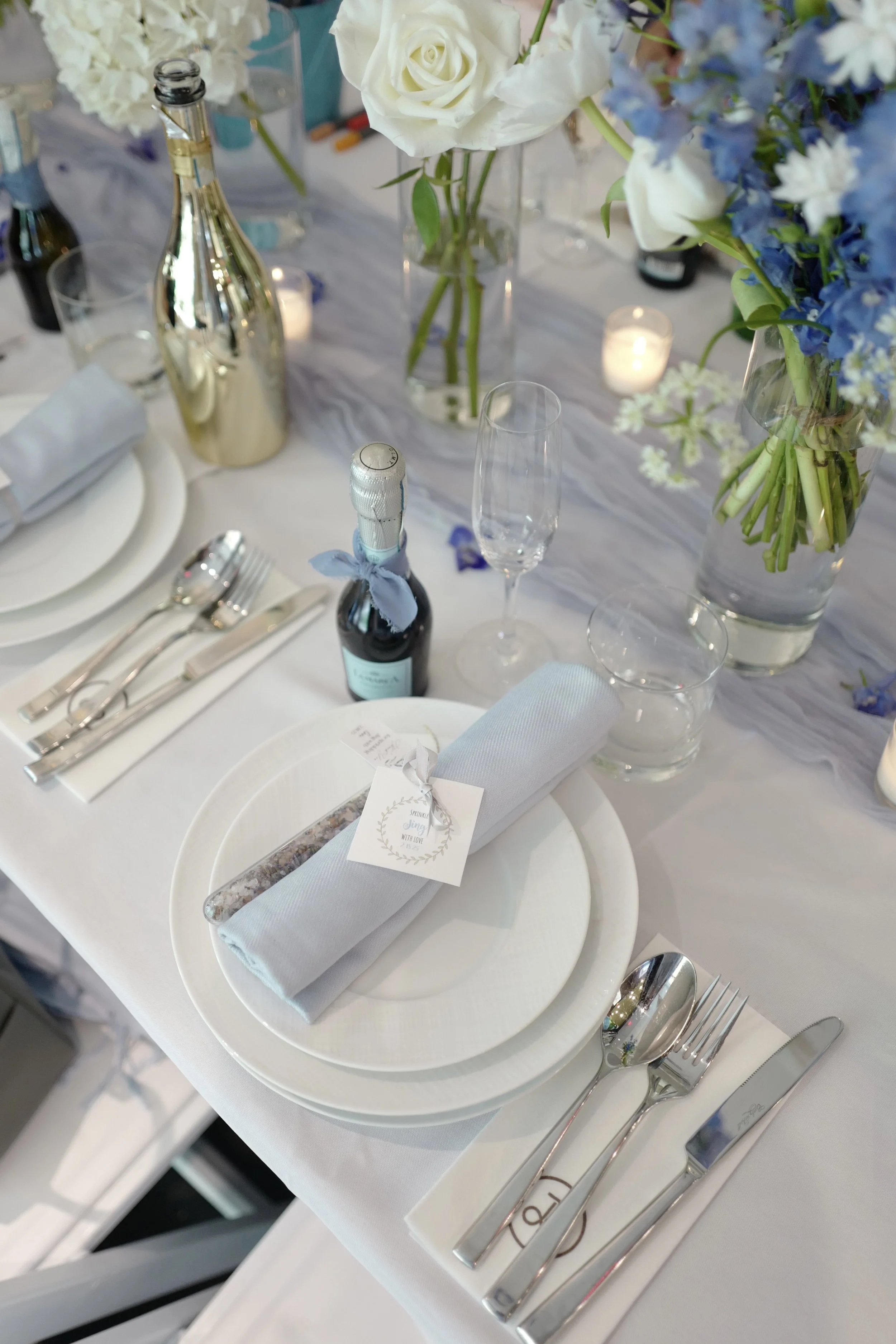 A decorated elegant table setting at a formal event, with white plates, silverware, wine glasses, a white napkin, a small favor or menu, and flower centerpieces with white and blue flowers, lanterns, and a bottle of champagne.