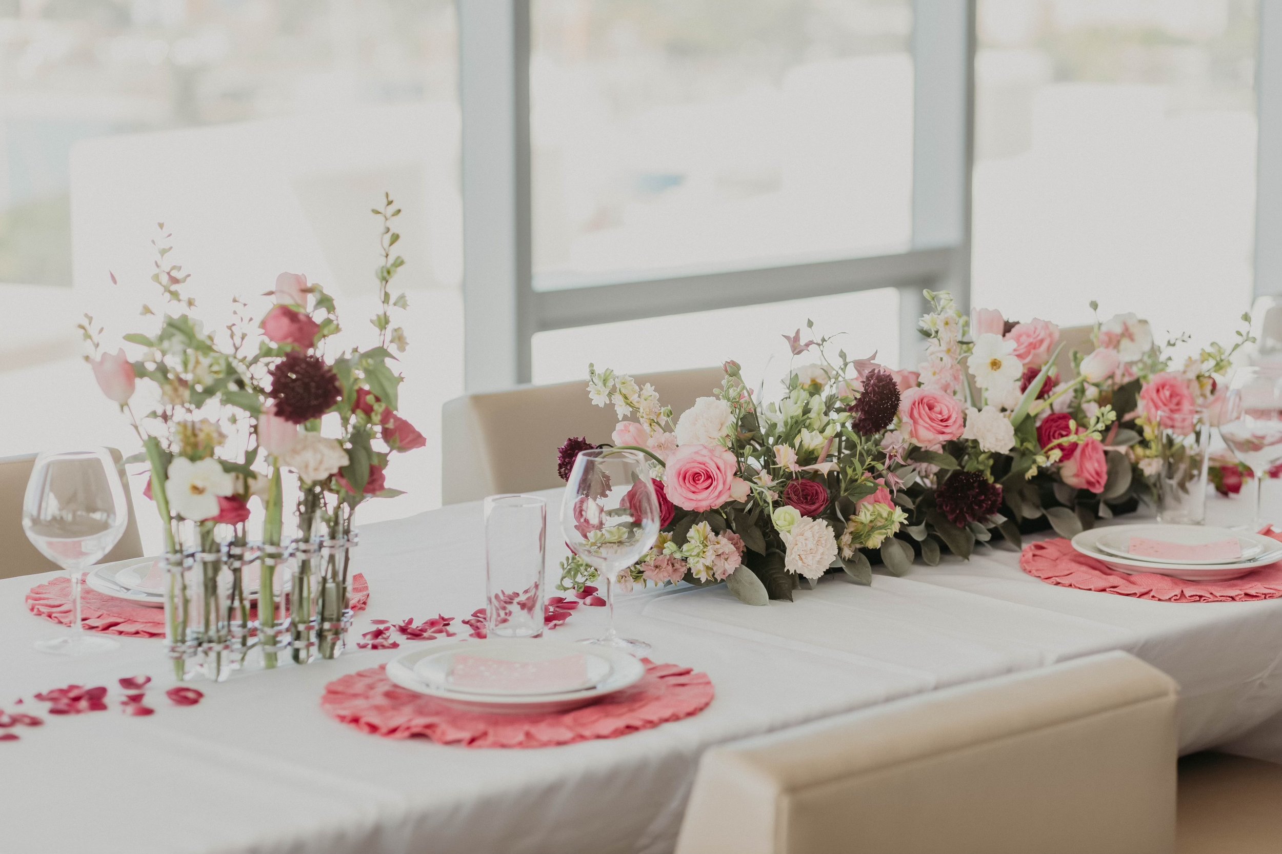 Elegant dining table decorated with pink and white floral arrangements, pink placemats, clear glassware, and white plates, set in a bright room with large windows.