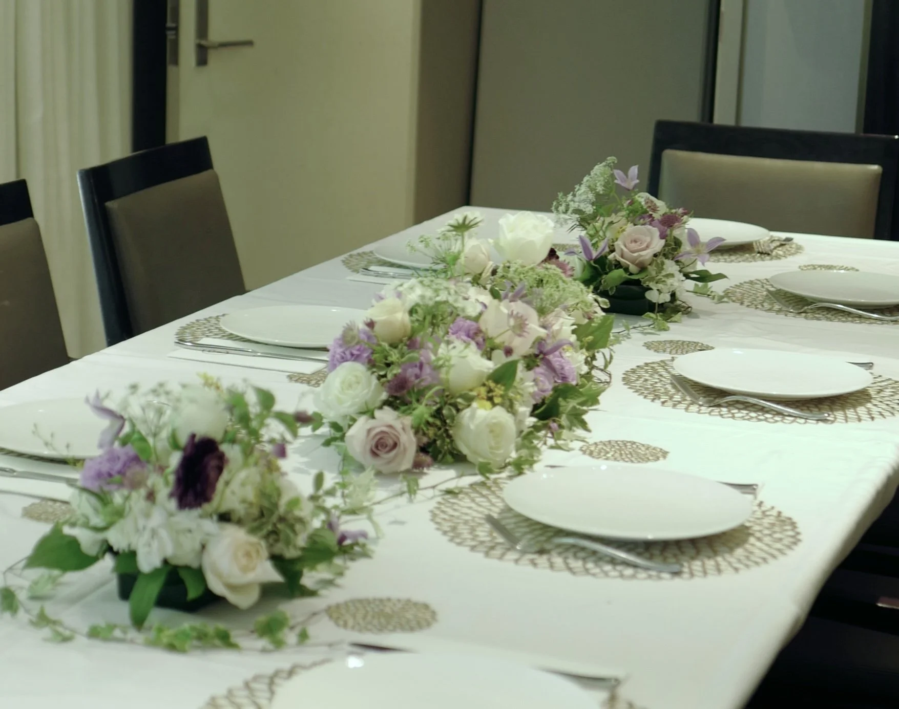 A dining table set with white plates, silverware, and decorative woven placemats. The table is decorated with three floral centerpieces consisting of white, lavender, and light purple flowers.
