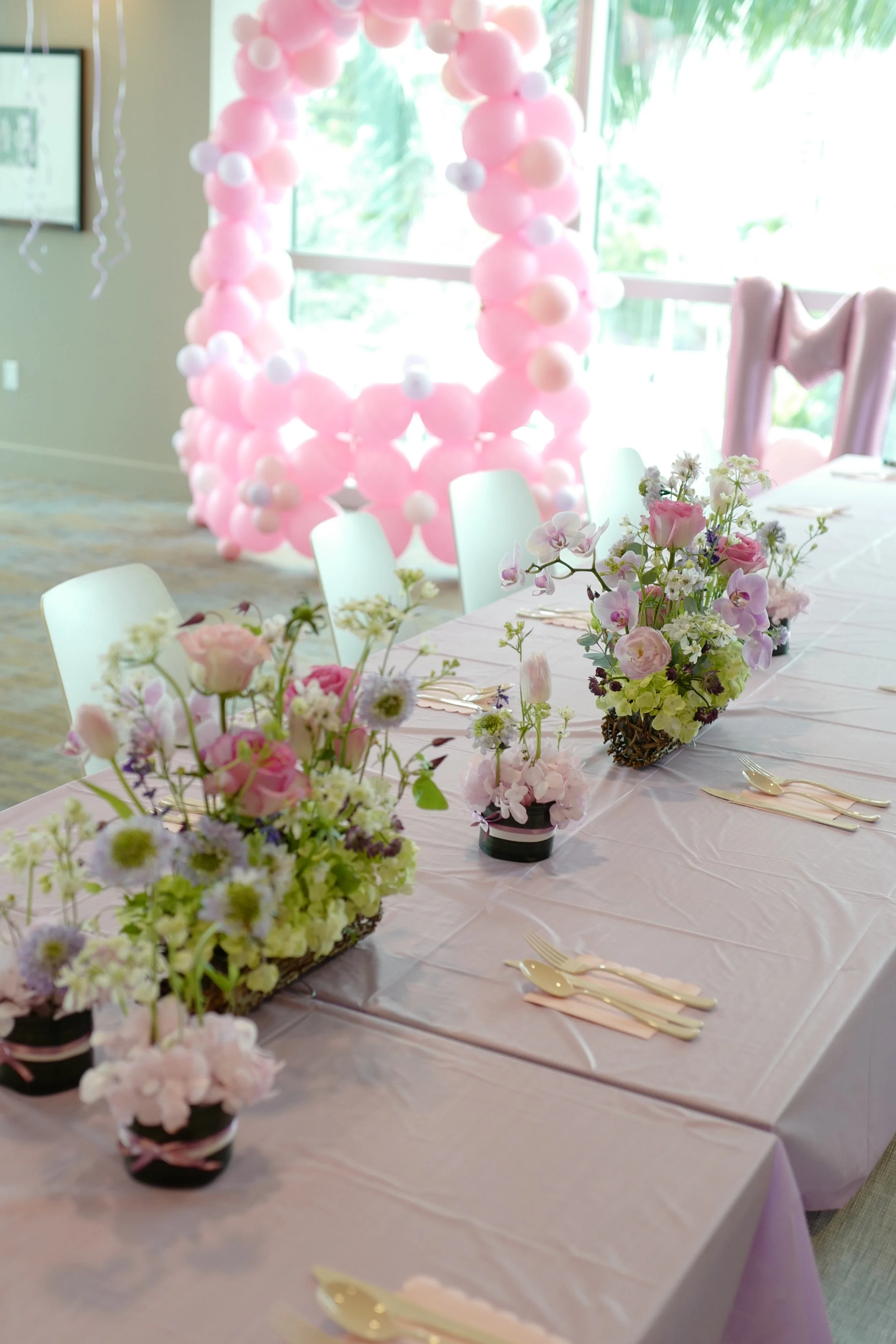 A decorated event table with pink and white floral arrangements, set for a celebration, with a backdrop of pink balloons.