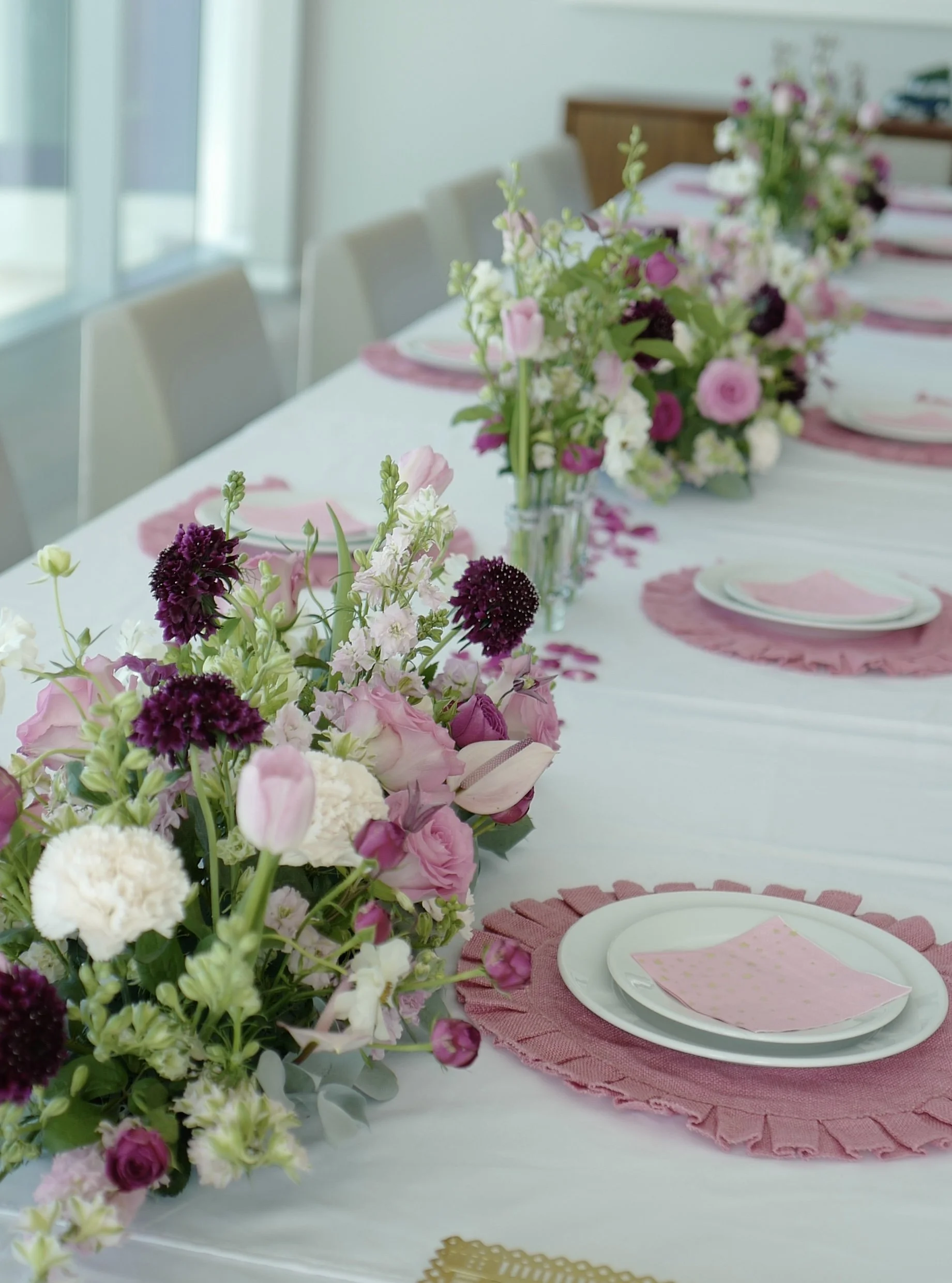 A long dining table decorated with pink and white floral arrangements, pink placemats, and white plates with pink napkins.