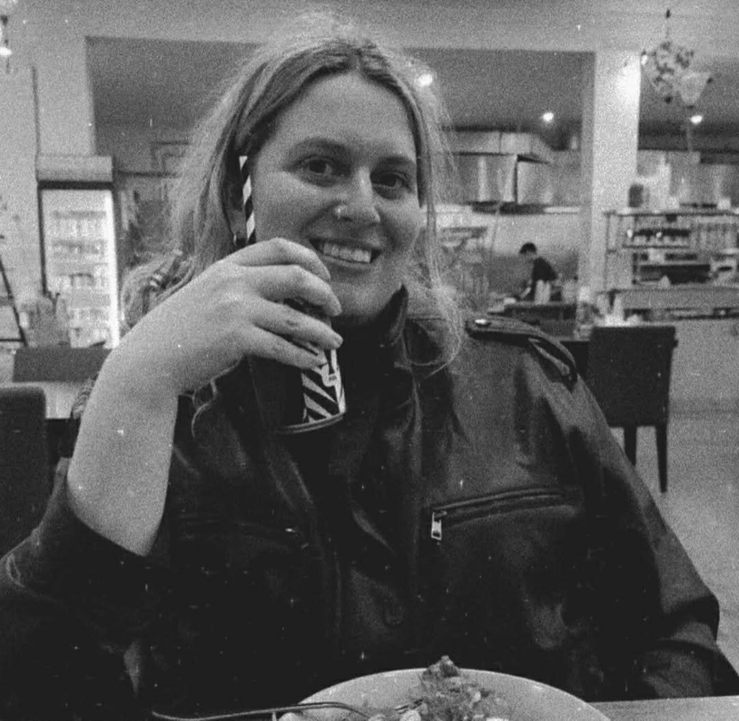 Black and white photo of a Eleanor smiling and holding a drink in a restaurant. She has light hair and is wearing a dark jacket, with a plate of food and utensils visible in front of her.