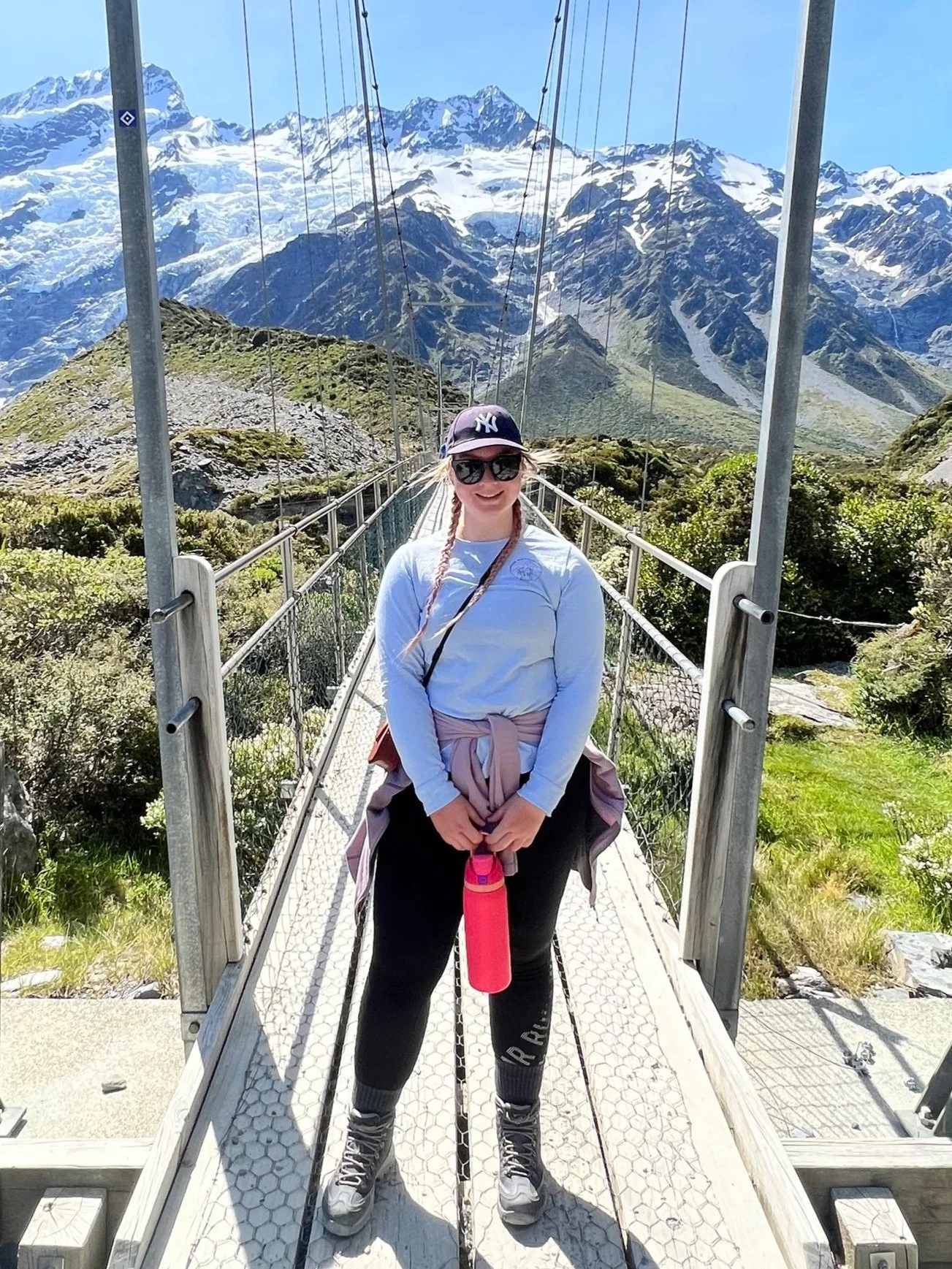 A woman standing on a suspension bridge in a mountainous area with snow-capped peaks in the background, wearing sunglasses, a baseball cap, a long-sleeve shirt, and hiking boots. Playing at Bailey and Zoeys Dog Daycare in Melbourne Altona North.