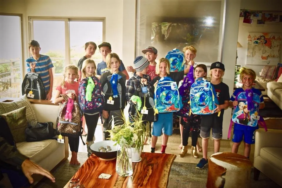 A group of children standing indoors holding backpacks and plush toys, with some smiling and posing for the camera.