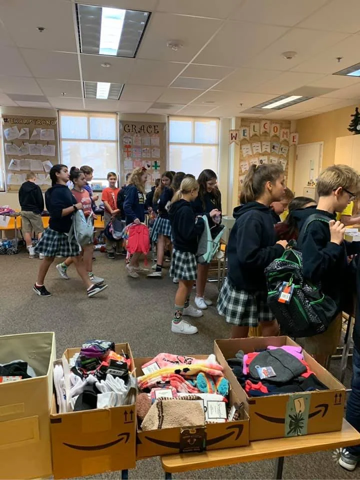A classroom with students lined up and waiting in line, wearing school uniforms consisting of plaid skirts and dark hoodies. There are boxes of supplies on a table in the foreground and posters on the walls, including one that says 'GRACE' and anothe