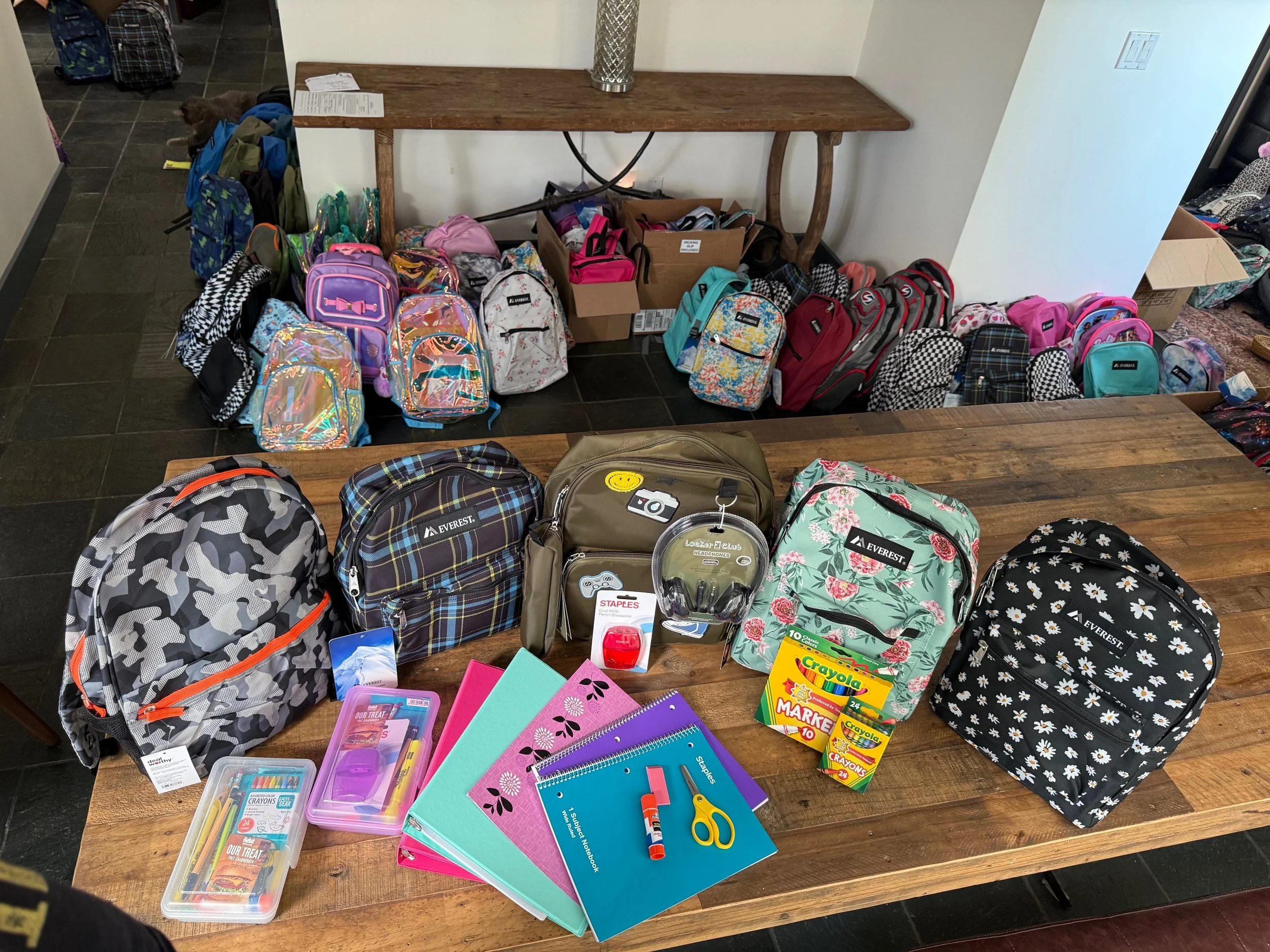 School supplies including backpacks, notebooks, scissors, glue, crayons, and boxes of crayons and markers are displayed on a wooden table, with more backpacks and bags on the floor and against the wall in the background.