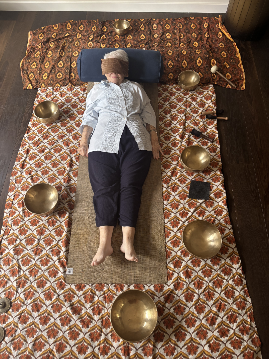 An elderly woman lying down on a patterned mat with a dark headrest, wearing a white shirt and black pants, with a blindfold covering her eyes. Surrounding her are six Tibetan singing bowls on a decorative cloth, and two mallets beside her right hand.