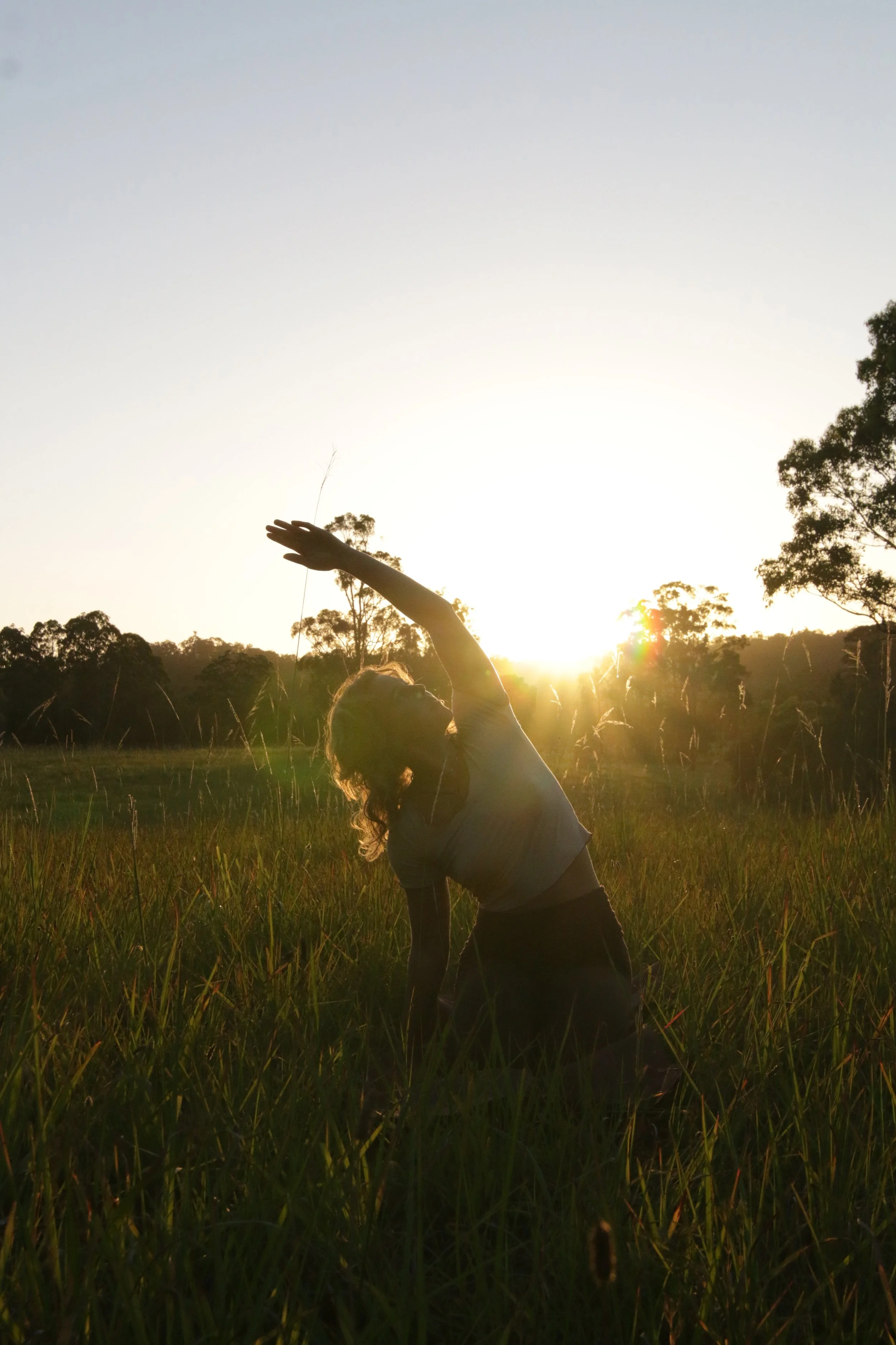 Person doing yoga or stretching in a grassy field during sunset with trees in the background.