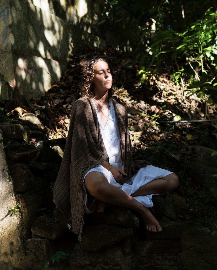 A woman practicing meditation outdoors in a natural setting, sitting cross-legged on rocks with her eyes closed, surrounded by trees and foliage.