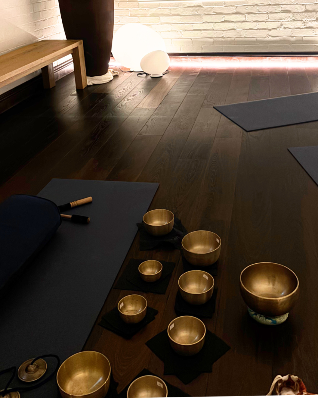 Yoga mats, meditation bowls, and gong on a dark wooden floor in a dimly lit room with a white brick wall.