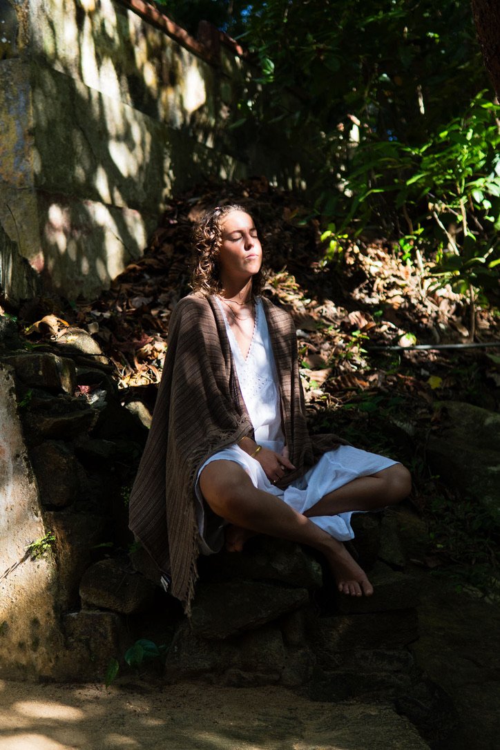 A woman sitting cross-legged and meditating outdoors in a wooded area with sunlight filtering through the trees.