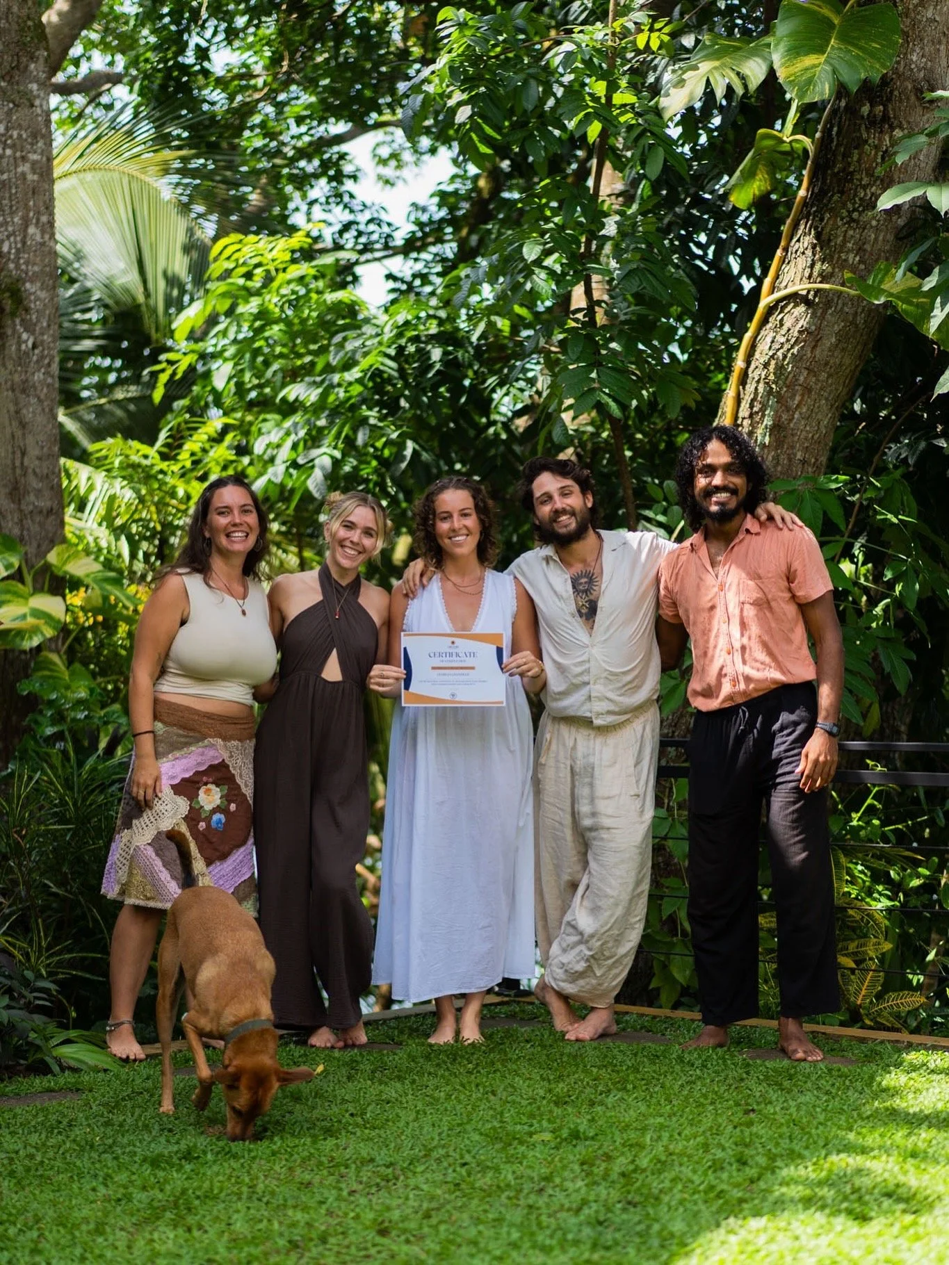 Group of five smiling people standing outdoors in a lush green garden, with three women and two men, one holding a certificate and a dog in the foreground.