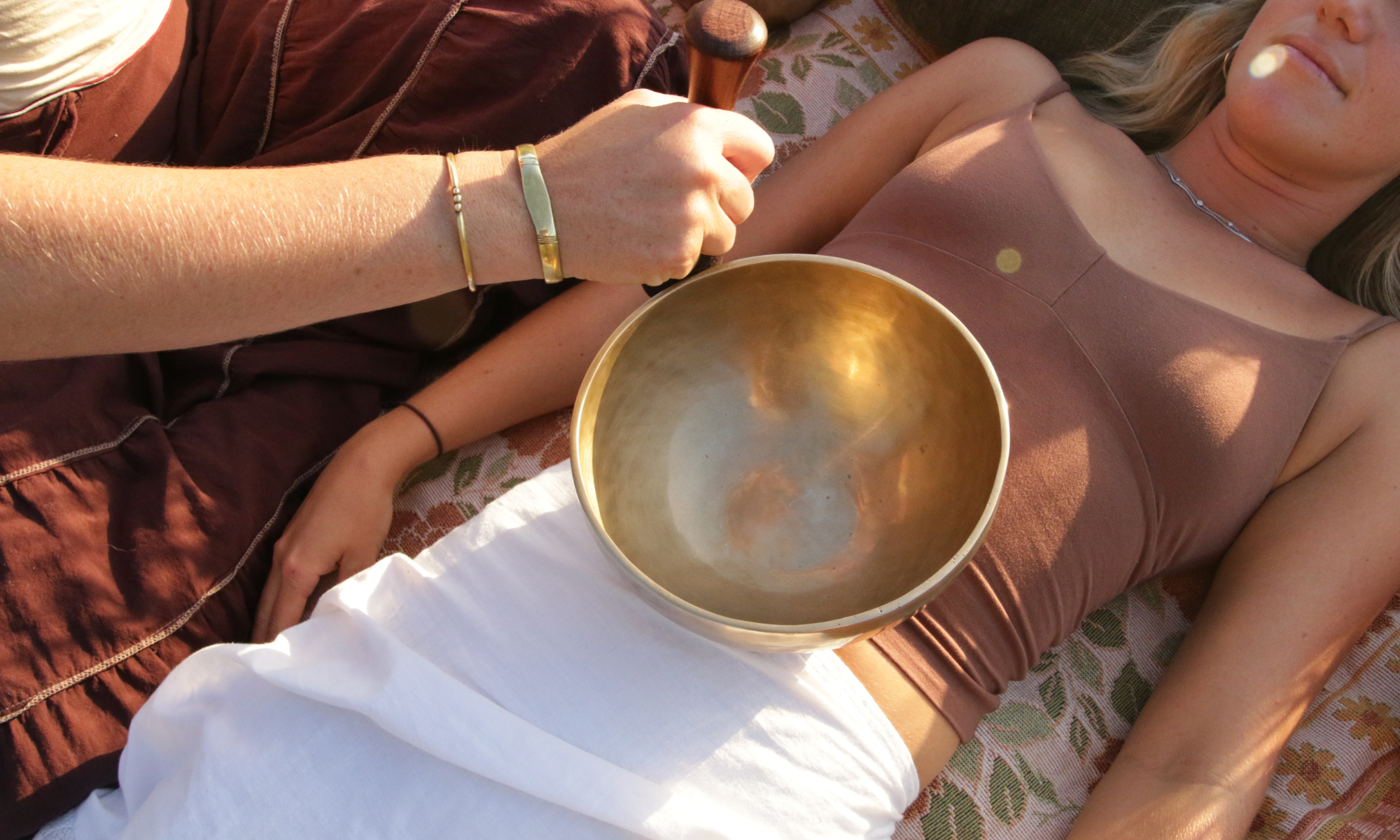 A person lying on a floral patterned blanket outdoors, holding a Tibetan singing bowl made of gold-colored metal, with another person next to them. The person holding the bowl is wearing a dark shirt, and the person lying down is wearing a muted pink dress, jewelry, and has blonde hair.