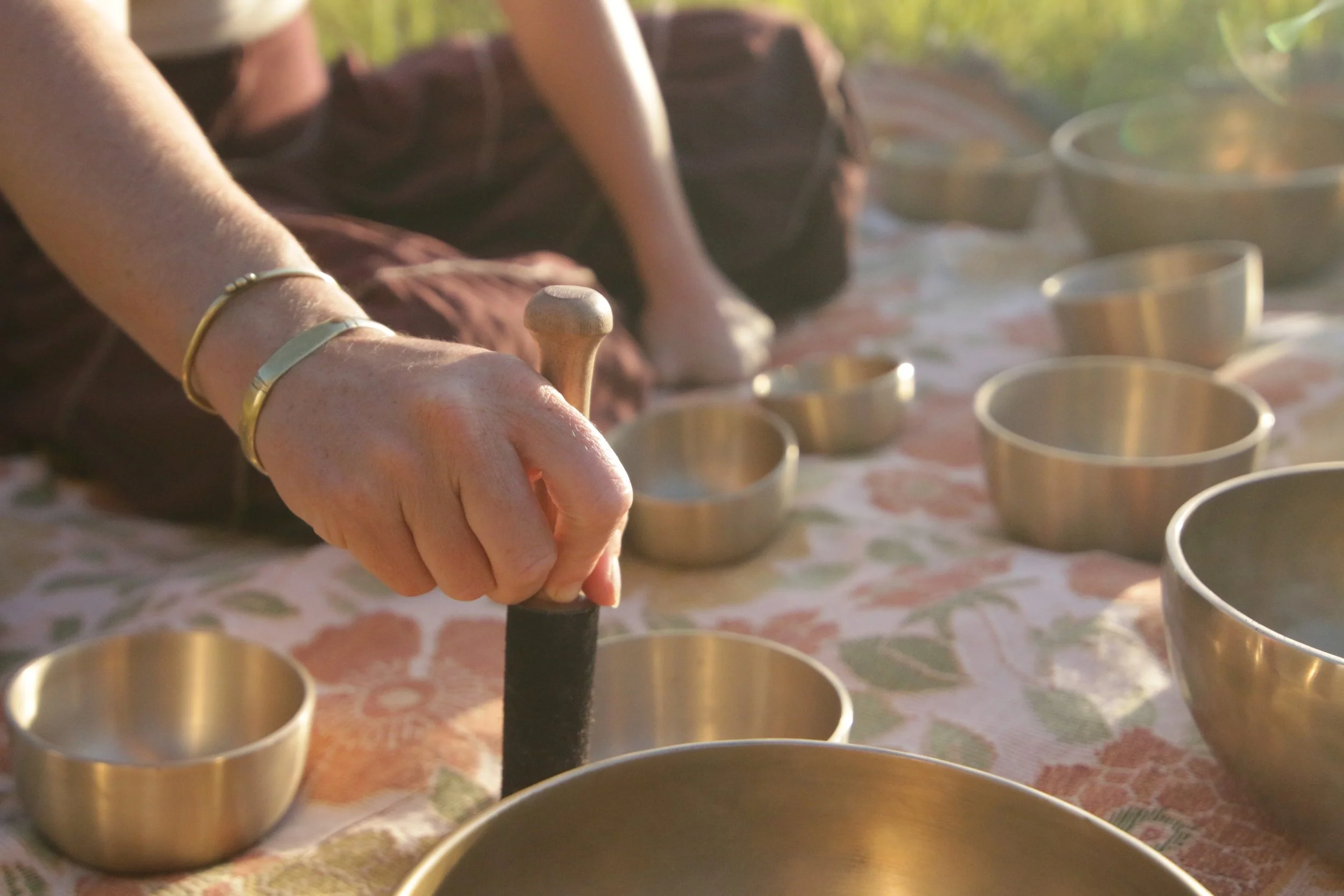Close-up of a person's hand pressing a black striker against the bottom of a metal lining in a small golden singing bowl, with more similar bowls arranged on a table covered with patterned fabric, outdoors in natural light.