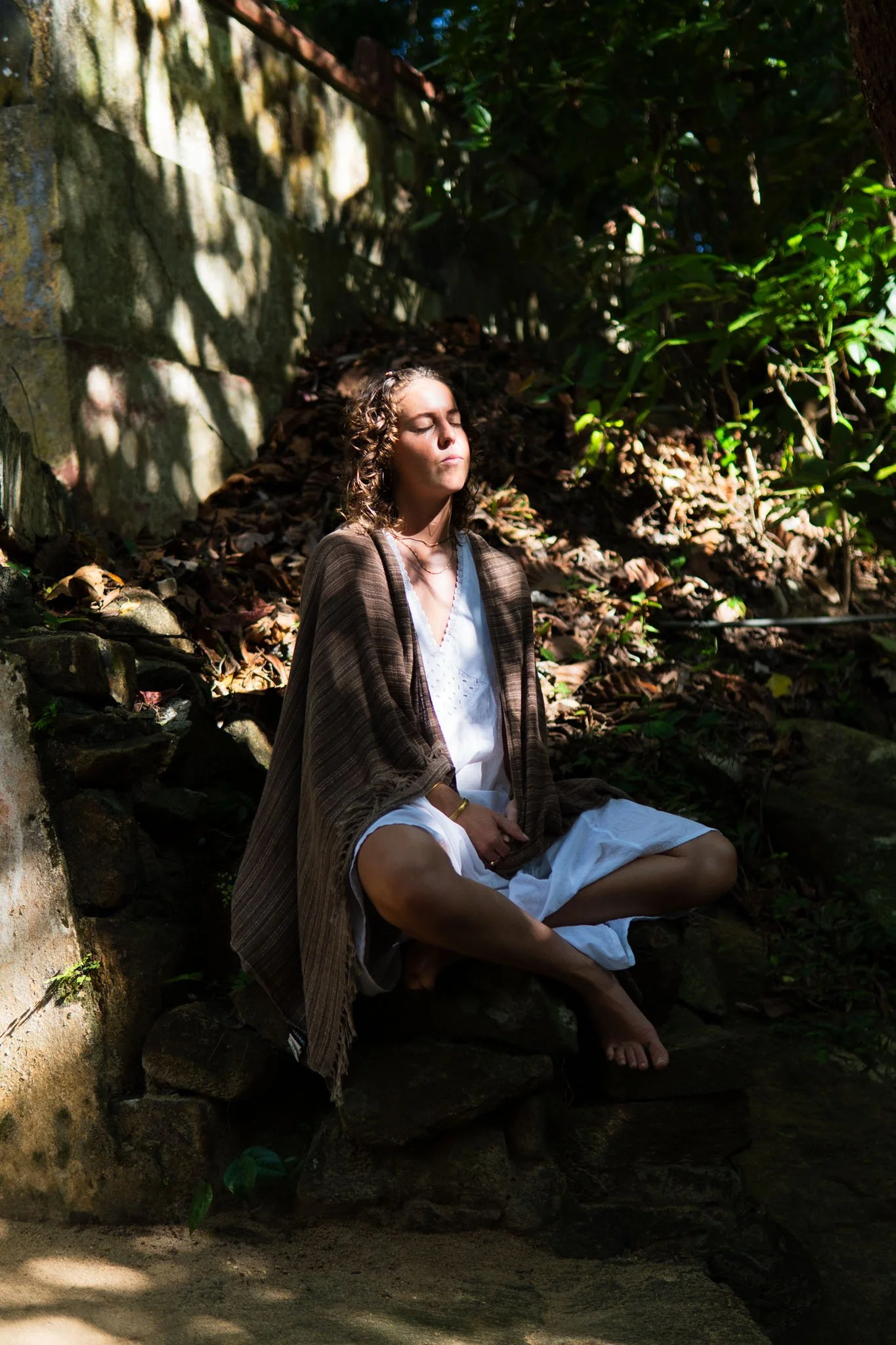 A young woman with dark curly hair sitting cross-legged on a rock, meditating with her eyes closed in an outdoor natural setting surrounded by greenery and shadows.