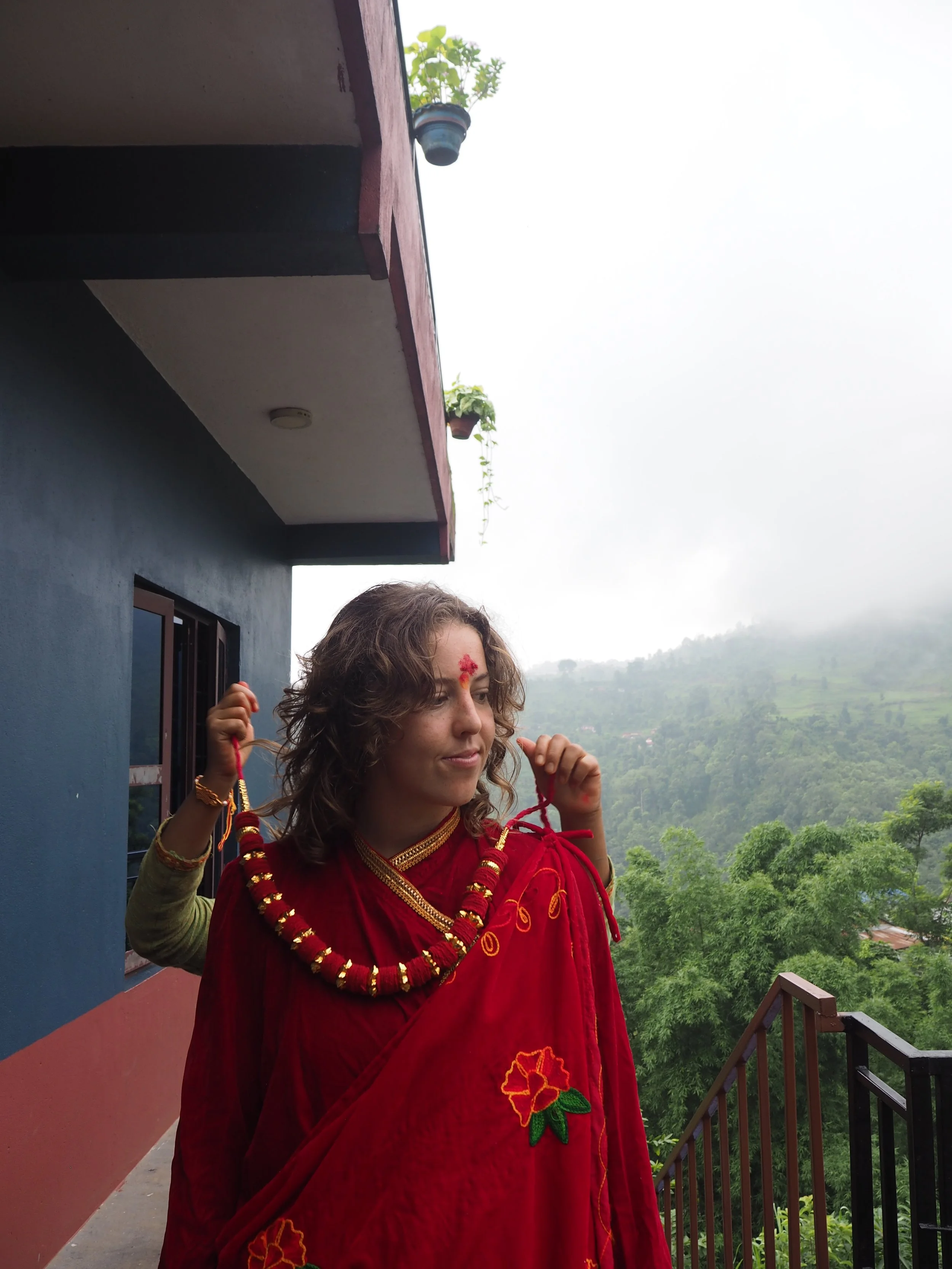 A woman dressed in traditional red attire with floral embroidery, standing on a balcony with lush green hills in the background, and a small part of a young person's hand and head visible behind her.