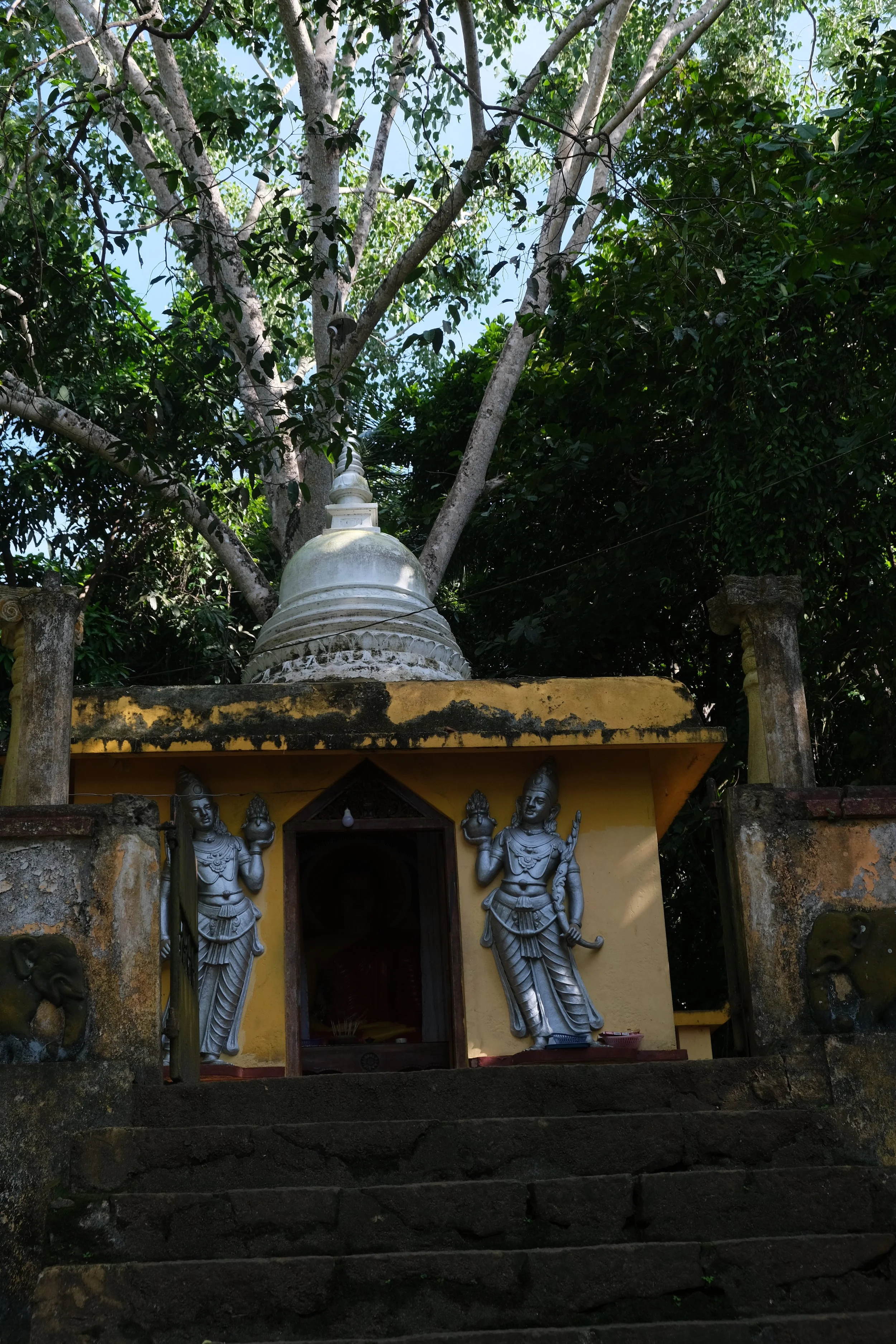 A small shrine with a yellow facade, featuring two silver statues of Hindu deities on either side of a doorway, with an ornate white stupa behind it. The shrine is surrounded by stone steps and a stone wall, with a large tree with green leaves in the background.