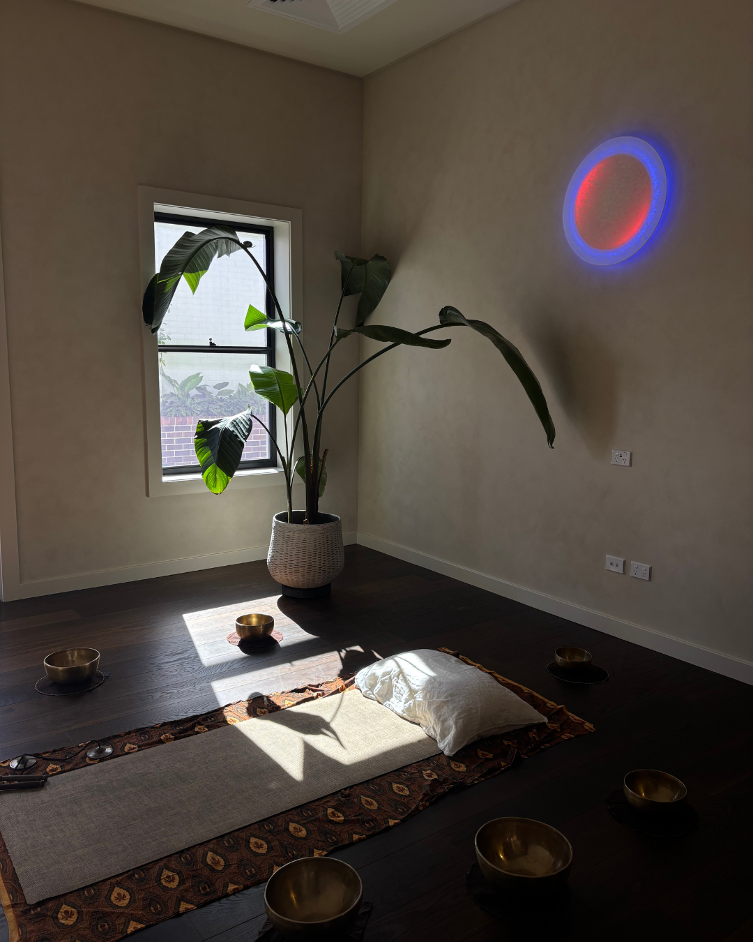 A peaceful meditation space with a beige wall, a large potted plant near a window, a small white rug with a pillow on top, surrounded by six brass bowls, a colorful patterned border around the rug, and a circular blue and red LED light on the wall.