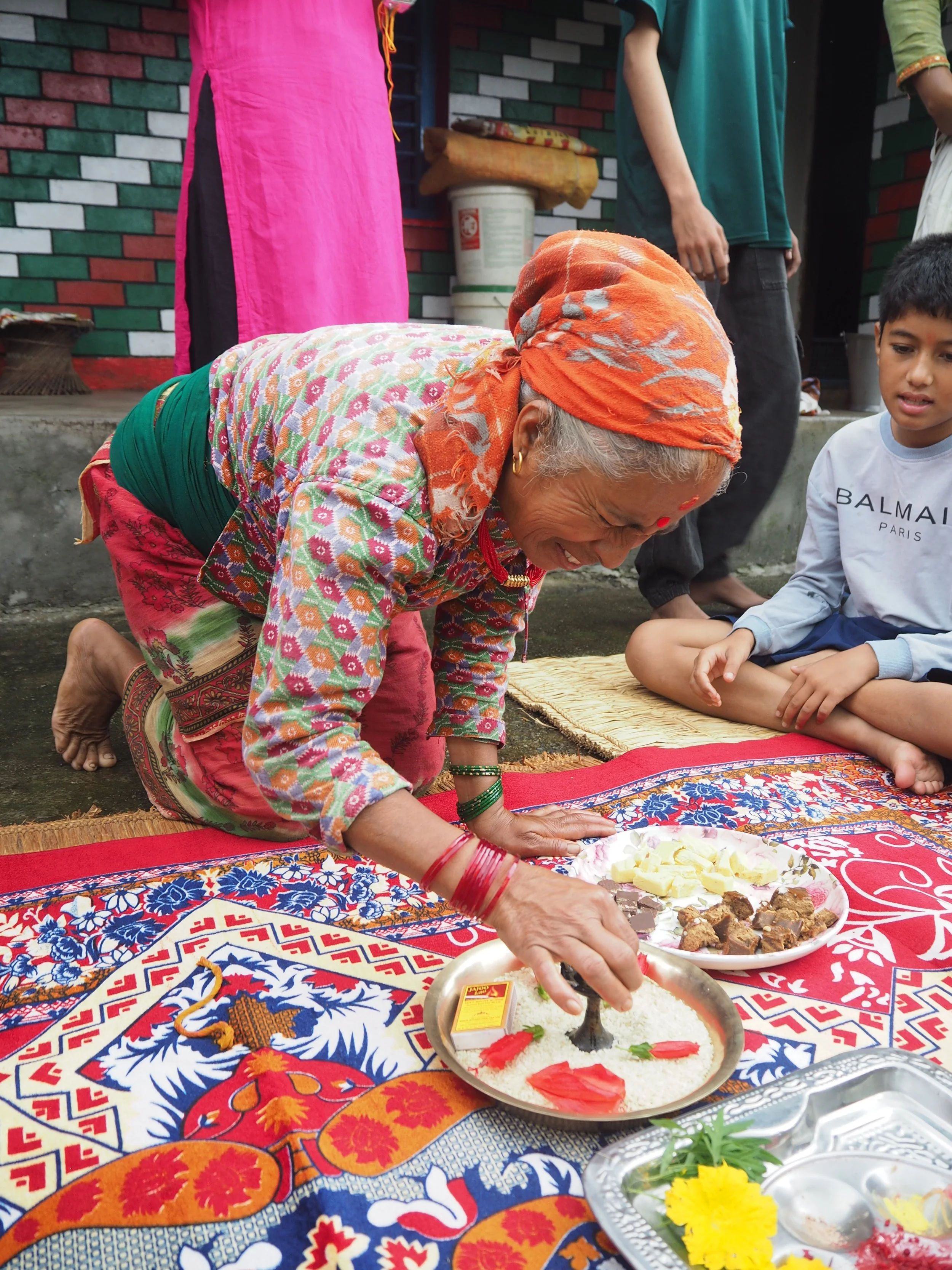 An elderly woman with gray hair, dressed in colorful traditional attire and headscarf, is kneeling on a carpet and serving food at an outdoor gathering. Several plates of sweets and rice are on the carpet, with one prominent plate having a black and white cake or sweet item. There are other people standing and a boy sitting nearby, participating in the event in front of a brick wall background.