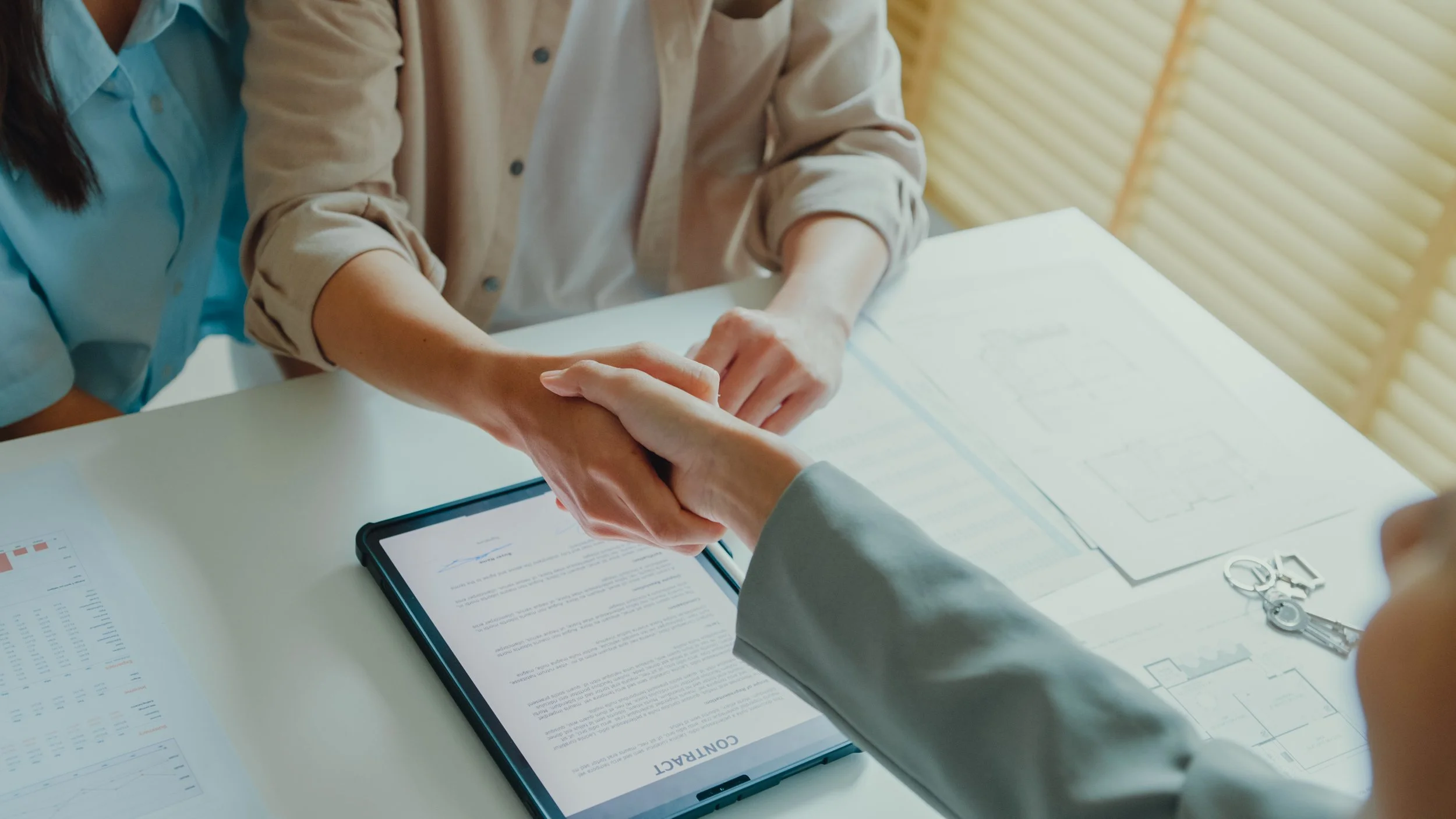 Two people shaking hands over a contract on a tablet on a desk, with documents and keys nearby.