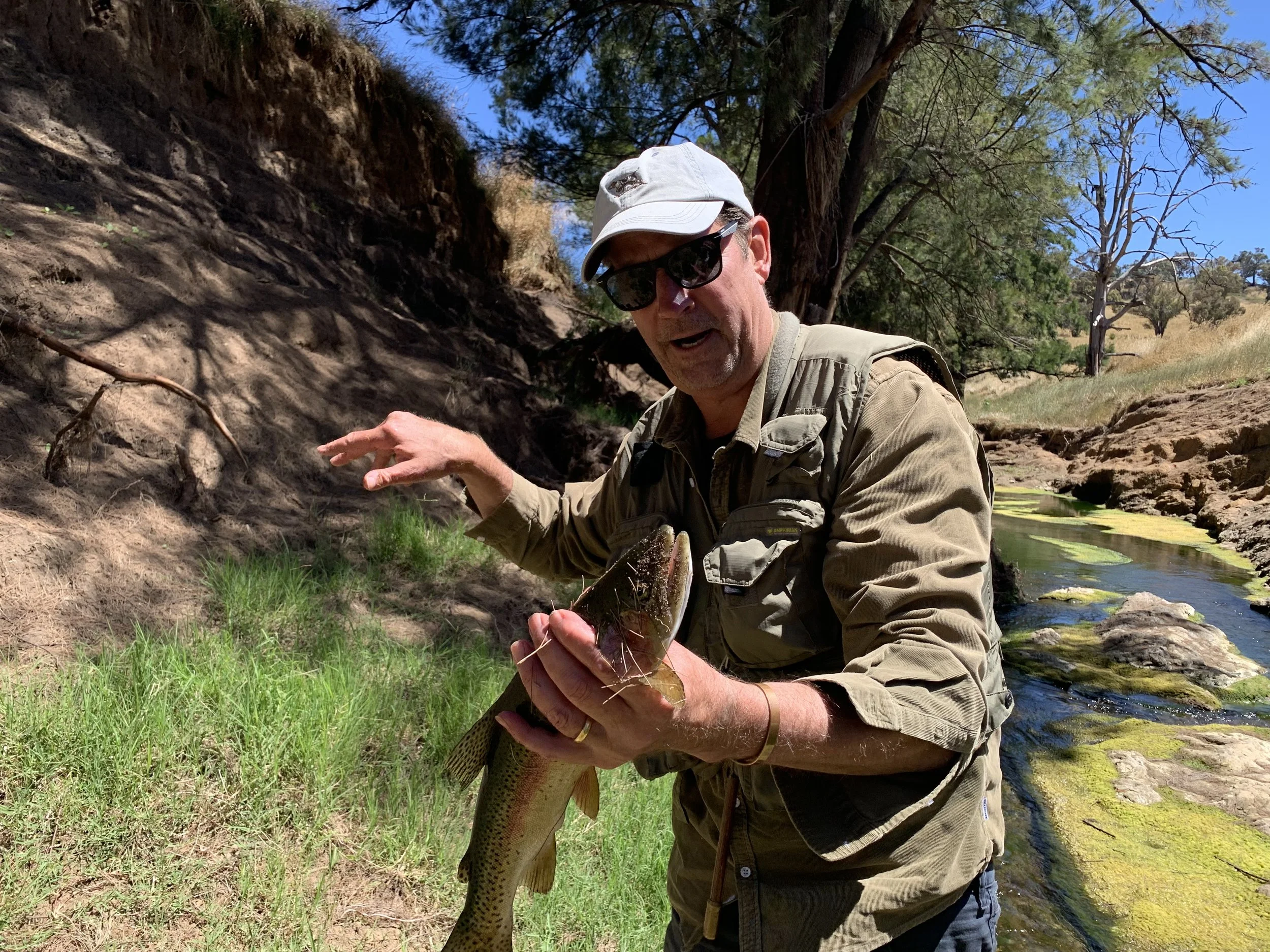 A man wearing sunglasses, a white cap, and a khaki jacket holding a large fish next to a creek in a natural outdoor setting.