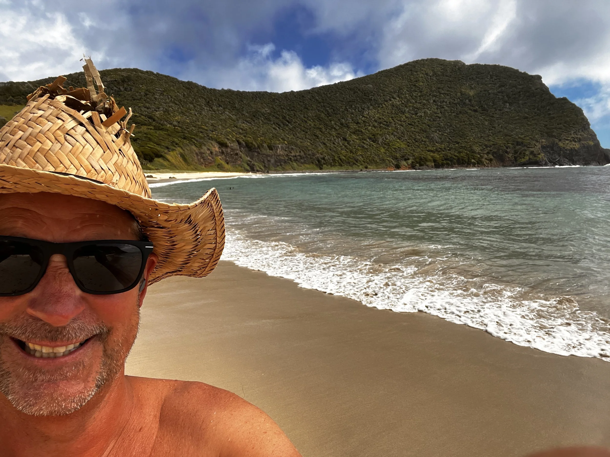 A man wearing a large straw hat and sunglasses taking a selfie on a sandy beach with a hilly, green landscape and ocean waves in the background.