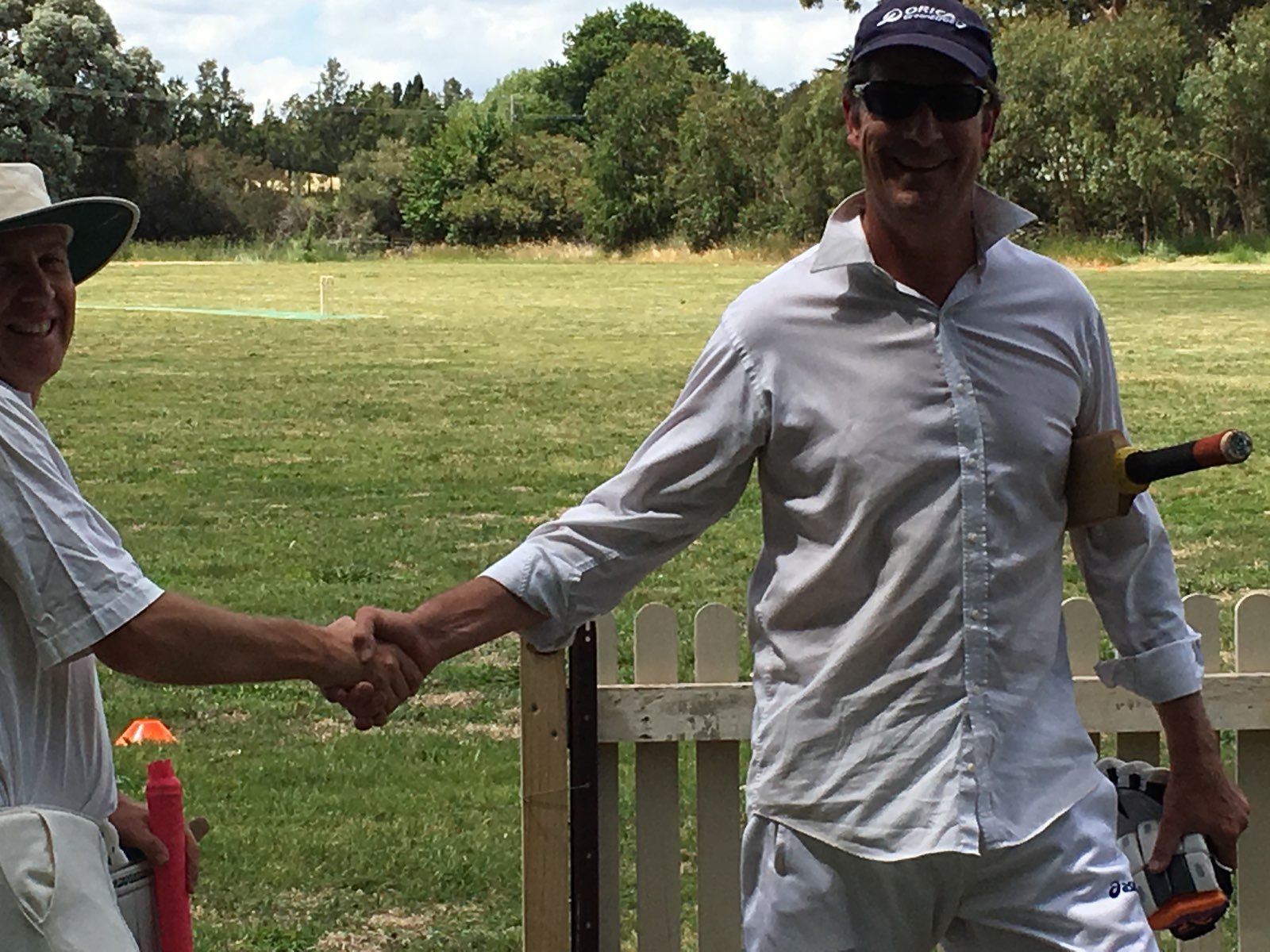 Two men in golf attire shaking hands on a golf course, with one holding a golf club and the other holding a pink golf tee or marker, and a wooden fence in the background.