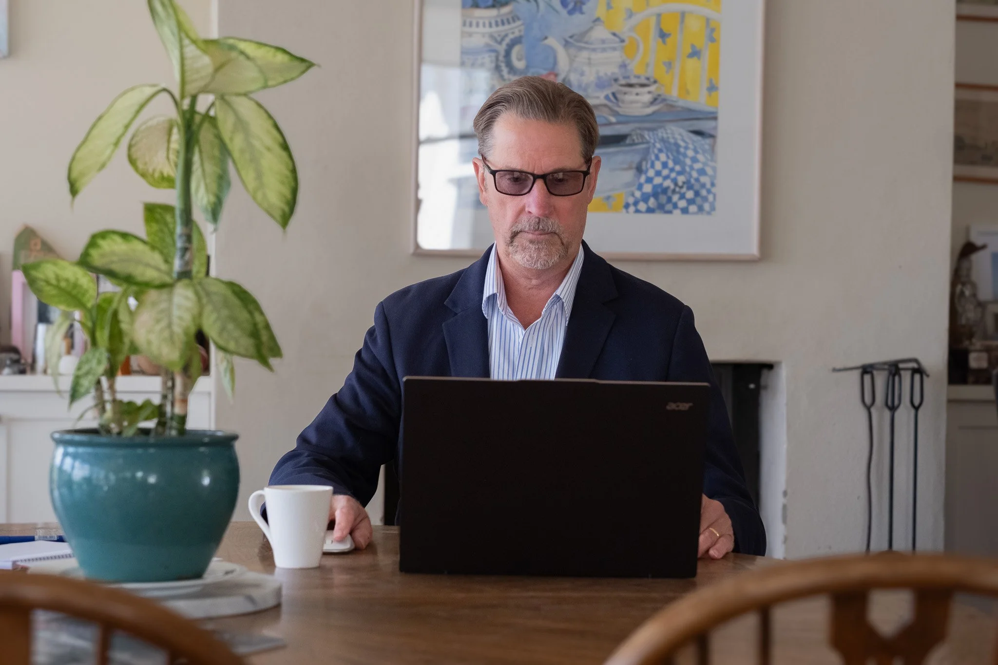 A man with glasses working on a laptop at a wooden dining table with a potted plant, a coffee mug, and papers.