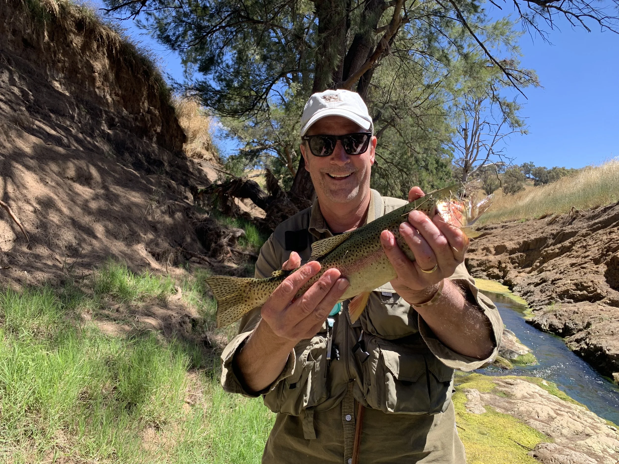A man wearing sunglasses, a white cap, and outdoor gear proudly holds a large rainbow trout he caught by a stream in a natural setting with green grass, trees, and rocky cliffs under a blue sky.