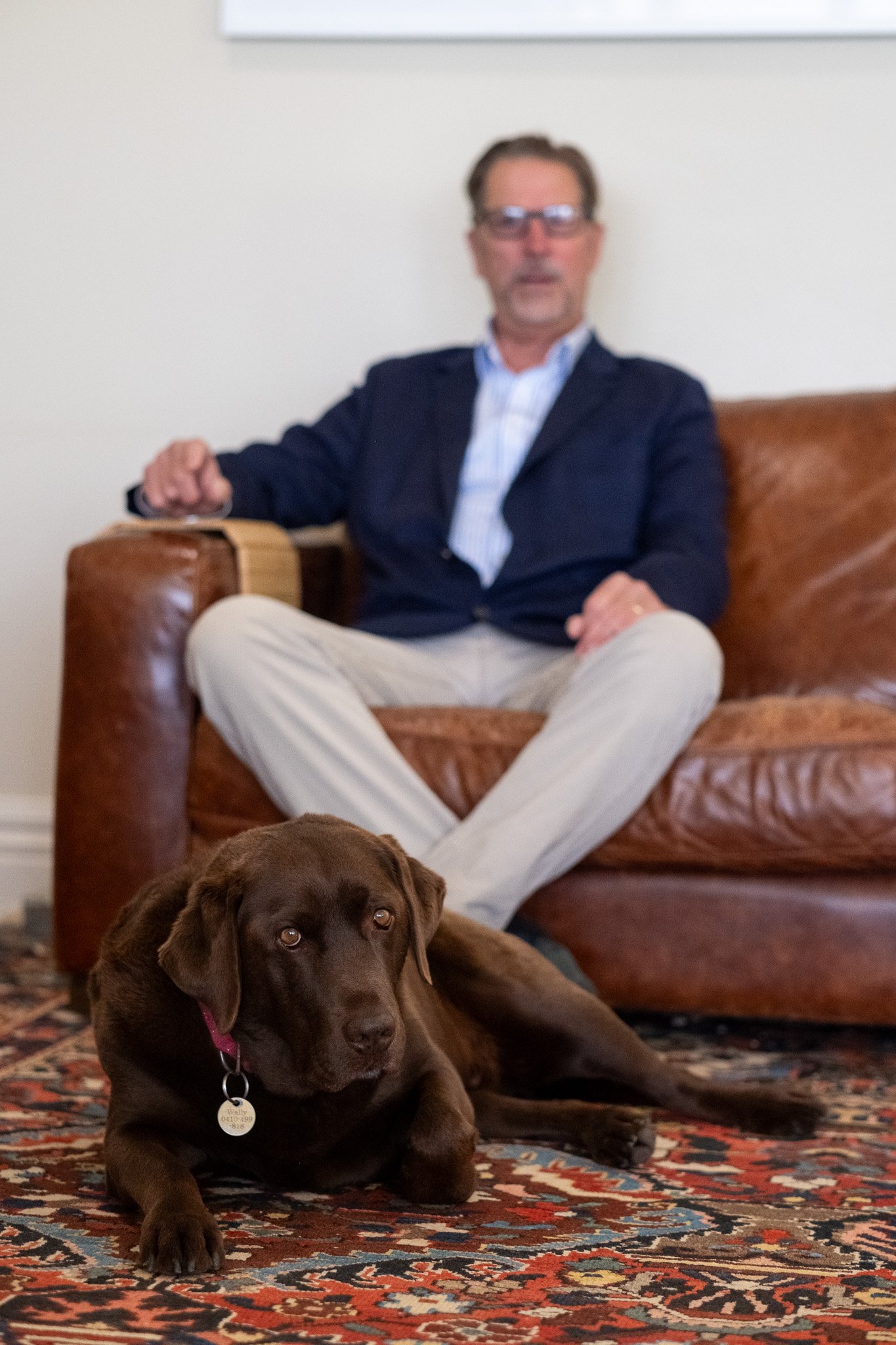 A man sitting on a leather couch with a chocolate Labrador retriever lying on an oriental rug in front of him.