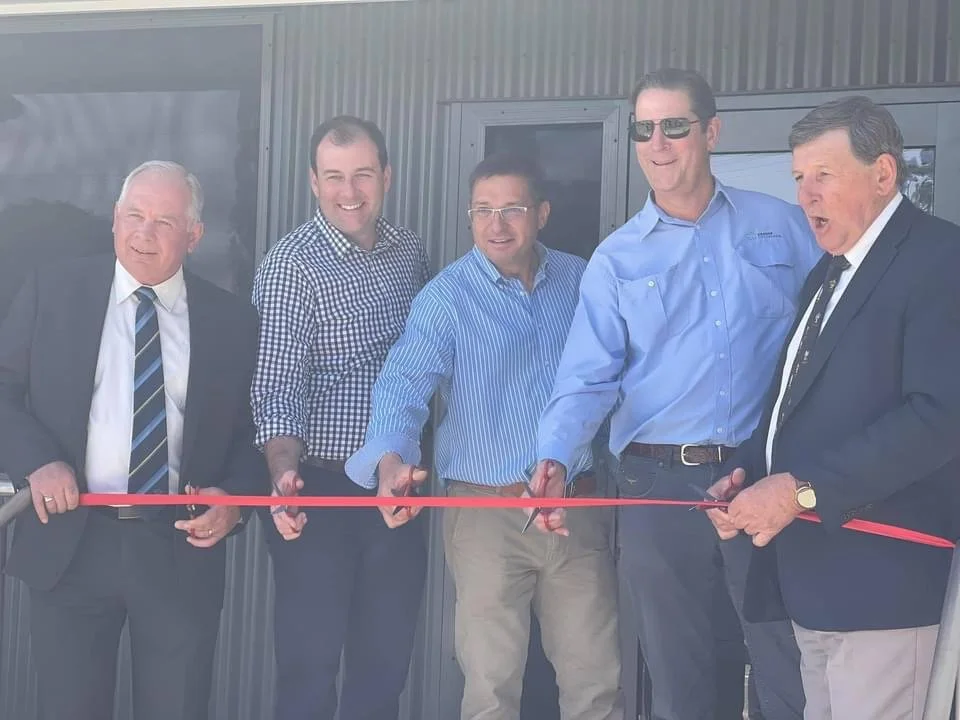 Six men standing outdoors in front of a modern building with glass and metal siding, participating in a ribbon-cutting ceremony. One man is cutting a red ribbon while others look on, smiling and clapping.