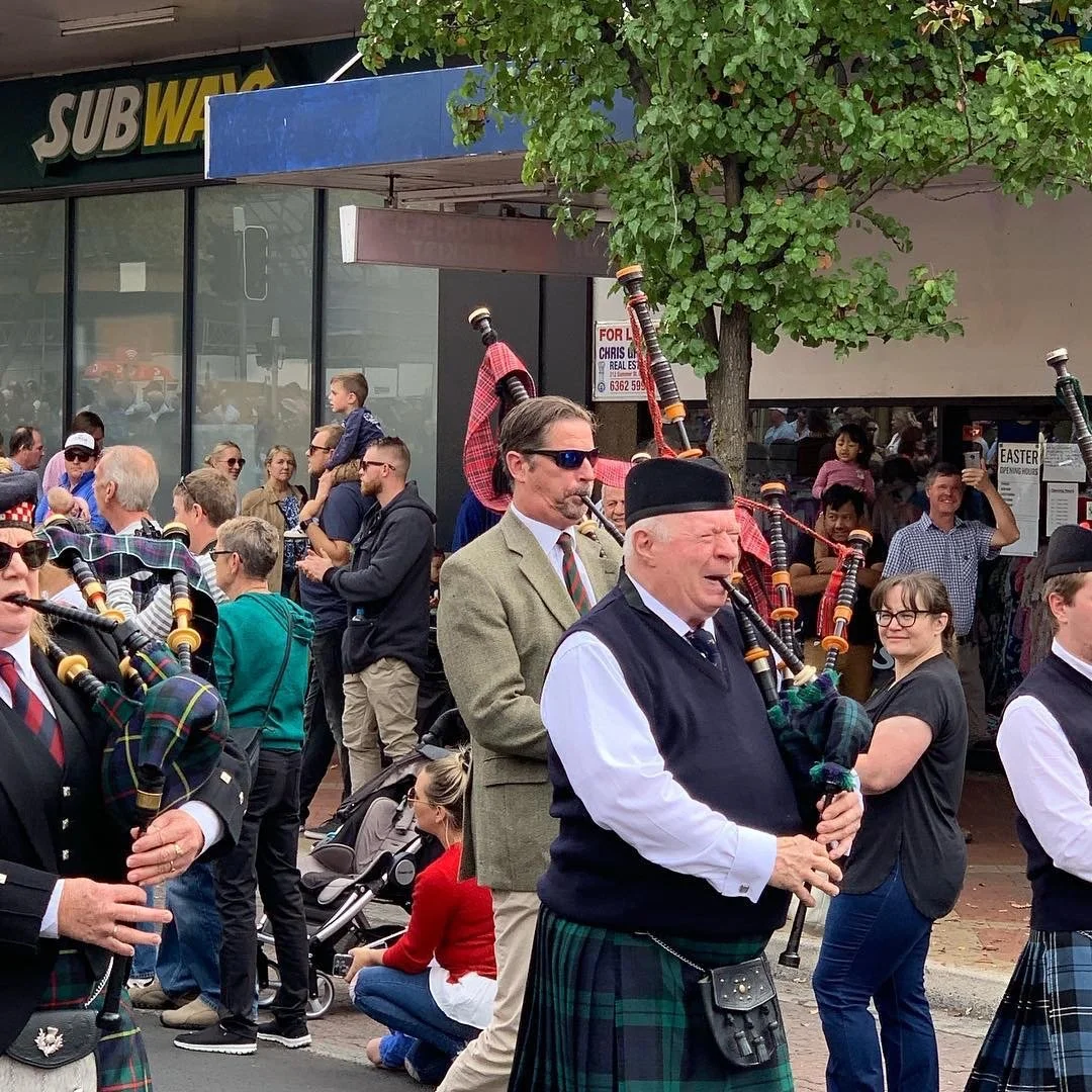 People marching in a parade, playing bagpipes, with a Subway store and a crowd of spectators in the background.