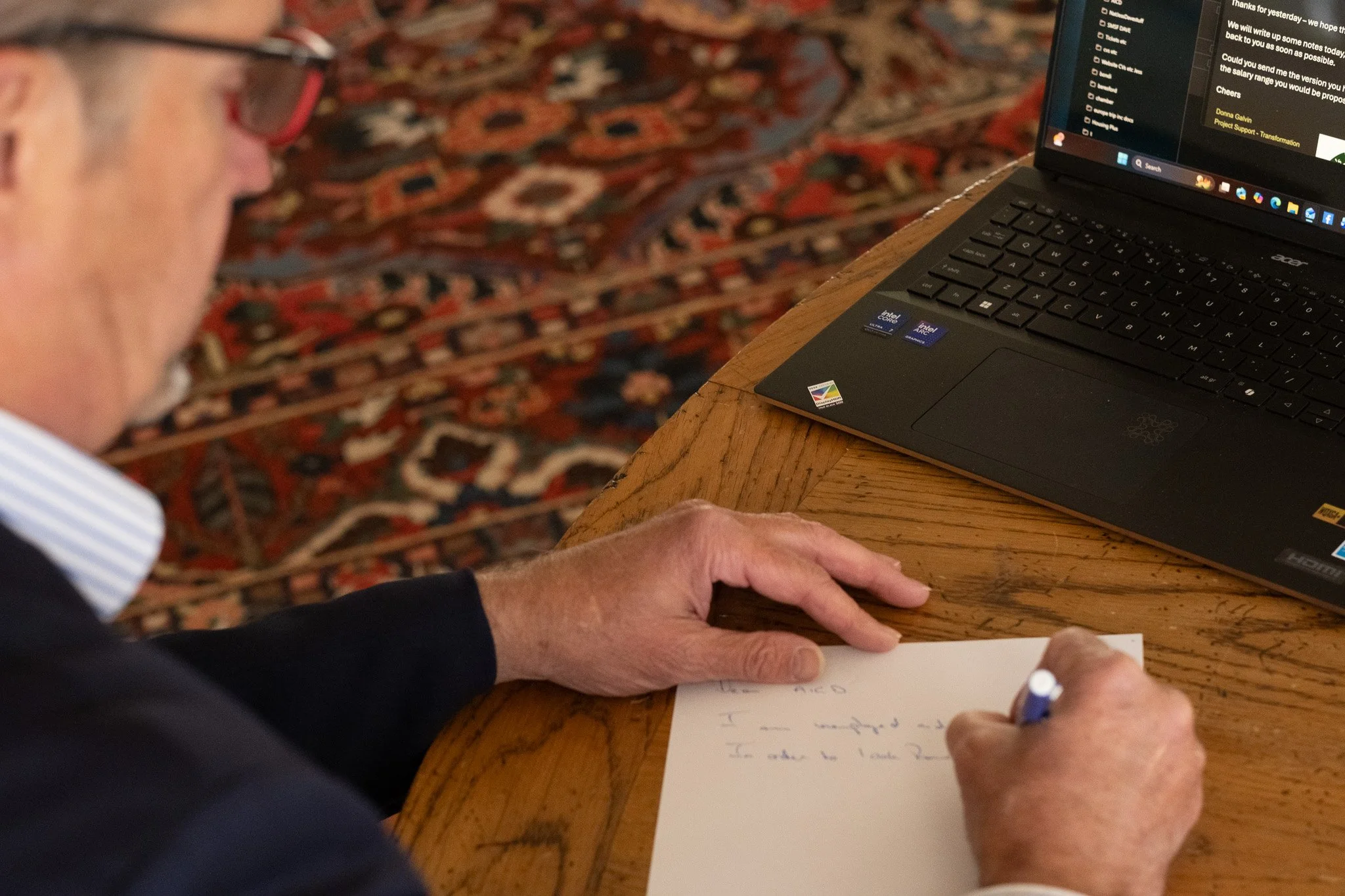 Older man with glasses writes on a piece of paper at a wooden table with a laptop nearby.